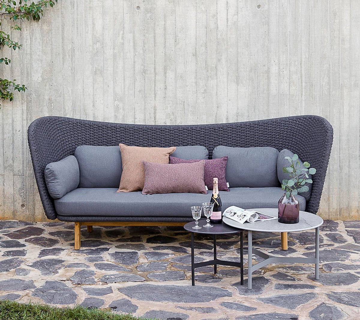 Un sofá moderno en gris con cojines decorativos, acompañado de una mesa pequeña con bebidas y un jarrón, situado contra una pared texturizada.