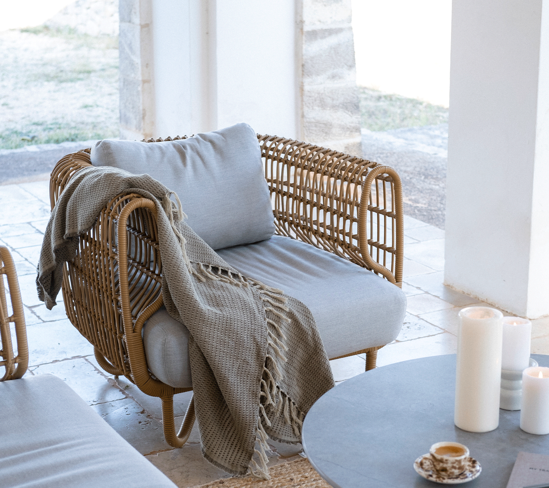 Silla moderna y acogedora con cojín gris claro, una manta de punto caída sobre el costado, rodeada de velas y una mesa minimalista.