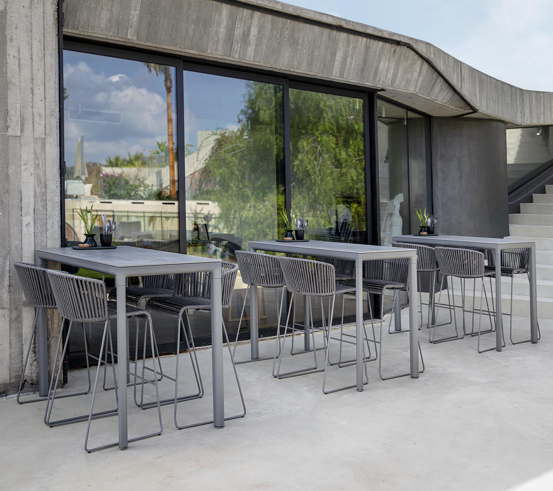Área de descanso moderna con mesas y sillas grises, con grandes ventanas que reflejan el verdor exterior. Estética de diseño minimalista.
