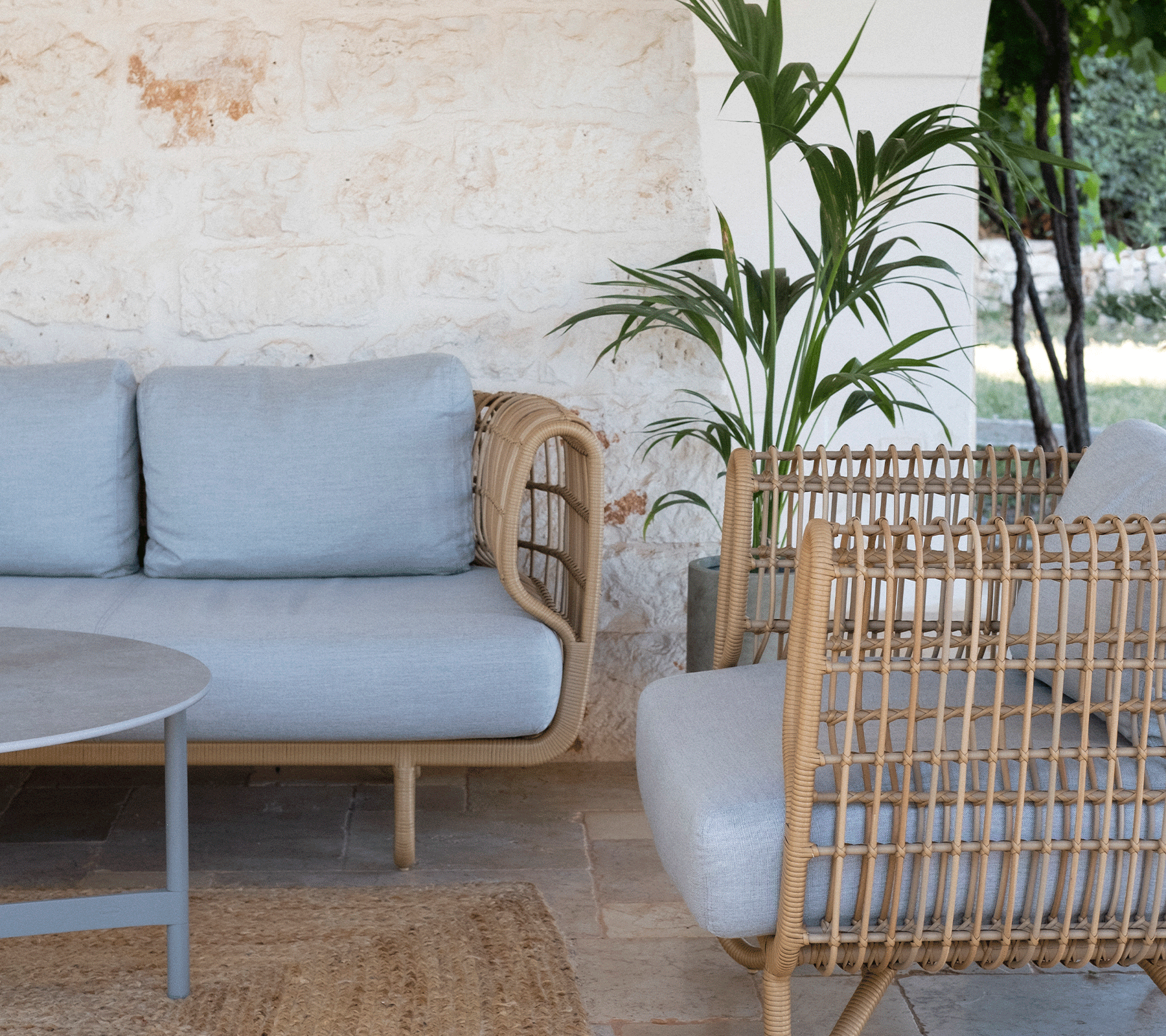 Área de descanso acogedora con un sofá gris claro, sillones de mimbre, una mesa redonda y vegetación de fondo. Adecuado para relajarse.