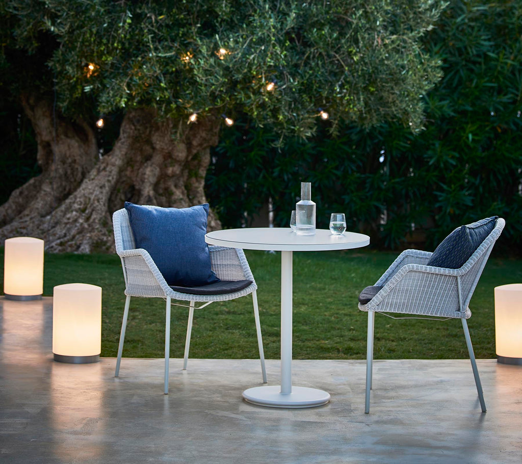 Un acogedor área de estar con dos sillas modernas, una mesa redonda y lámparas elegantes, rodeada de vegetación y luz cálida.