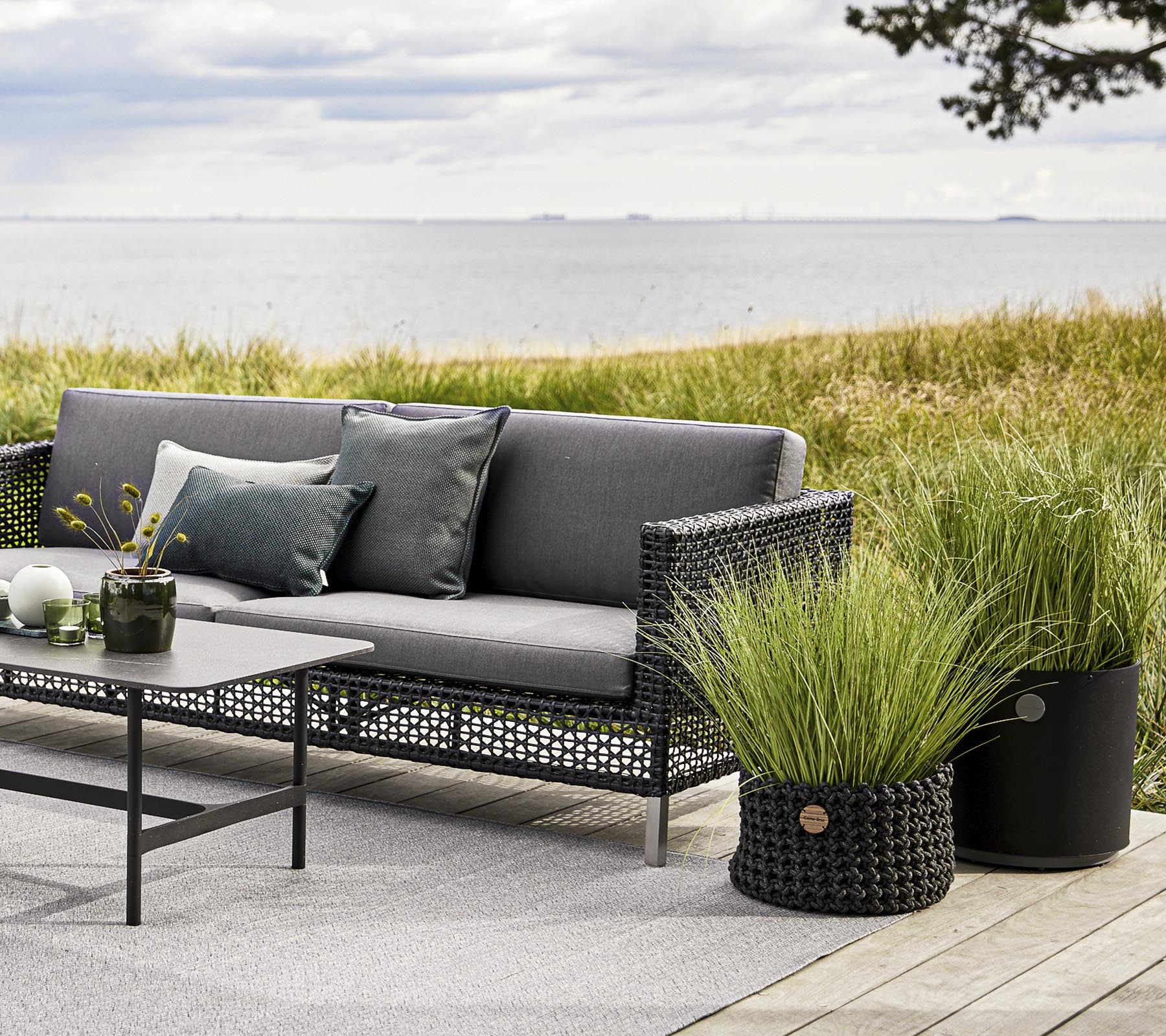Mobiliario exterior elegante con un sofá gris, cojines decorativos y una mesa, complementado con macetas decorativas de hierba y un fondo sereno.