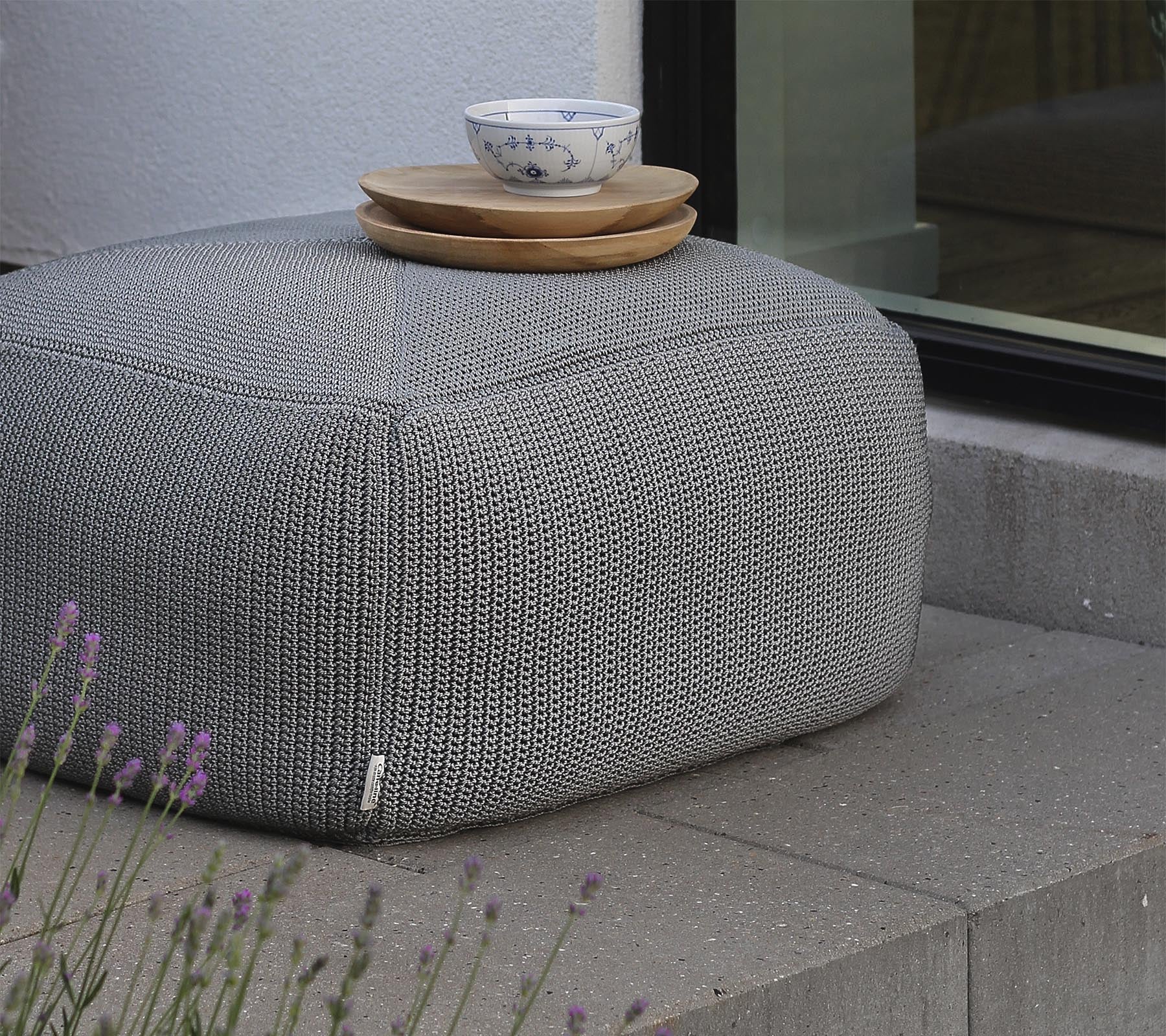Un puff gris texturizado cubierto con platos de madera y un tazón de cerámica, situado cerca de una ventana y rodeado de lavanda.