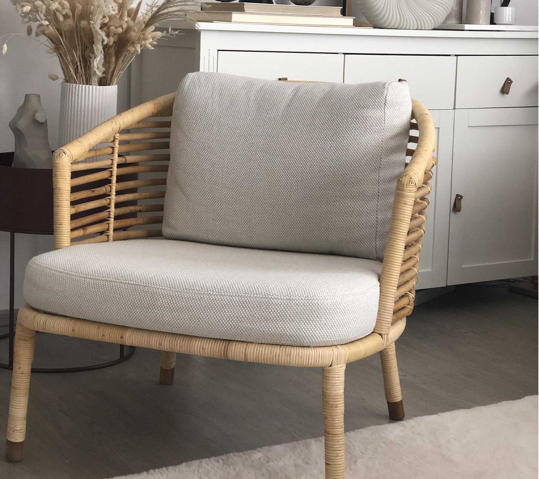Una silla elegante con un diseño tejido y cojines suaves, acompañada de una planta decorativa y una unidad de almacenamiento elegante en el fondo.