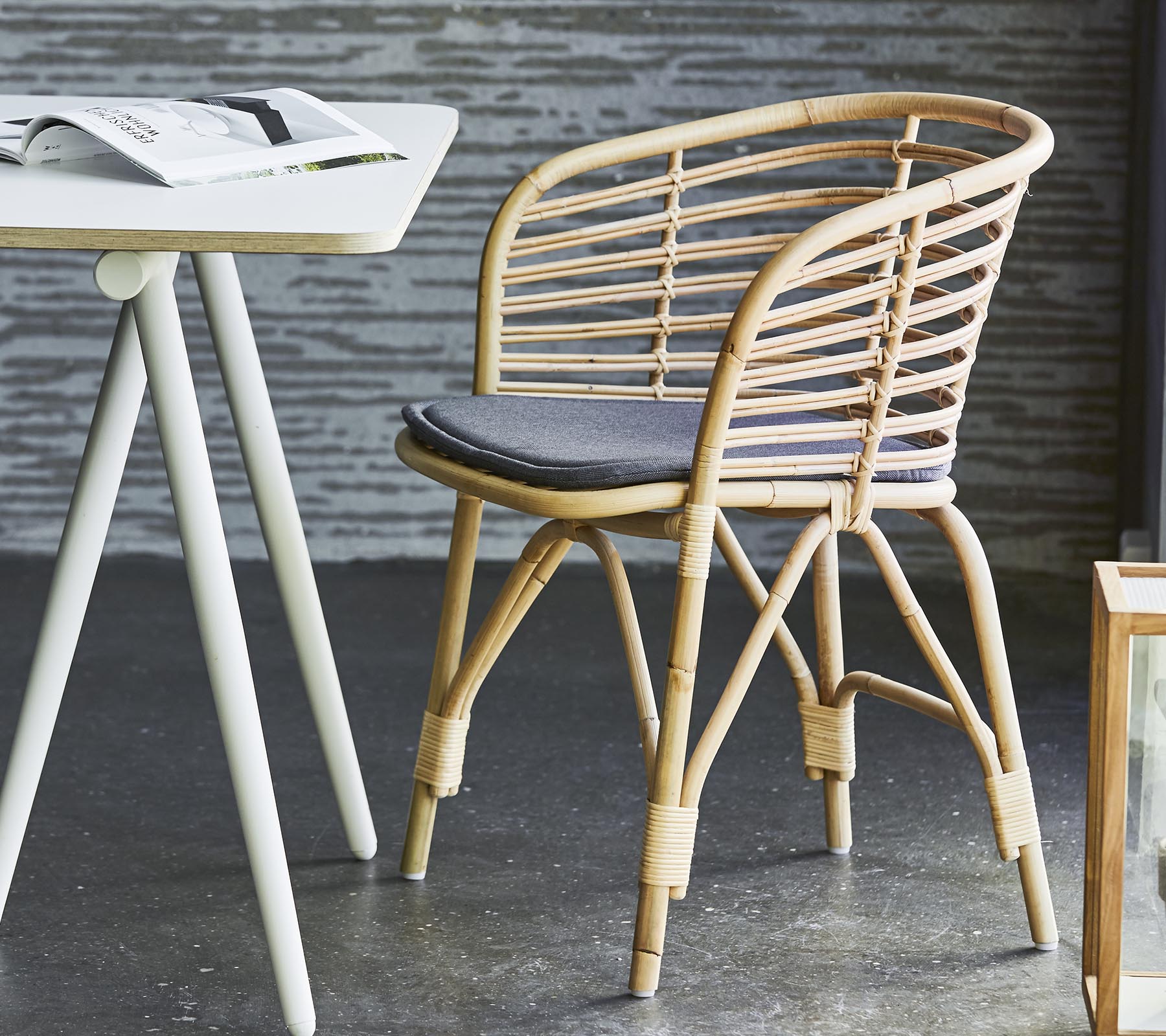 Silla de mimbre elegante con un cojín gris, combinada con una mesa simple y decoración contemporánea. Apta para asientos informales y relajación.