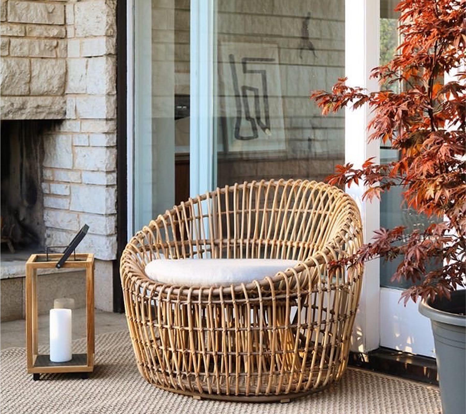 Una silla de mimbre elegante con un cojín, acompañada de una linterna y una planta vibrante, que realza un espacio acogedor e invitante.
