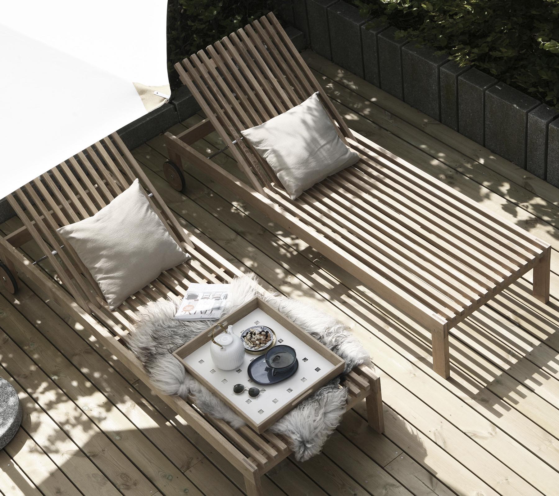Dos sillas de lounge de madera con cojines están dispuestas junto a una mesa pequeña adornada con una manta acogedora y artículos decorativos. La luz natural filtra a través.