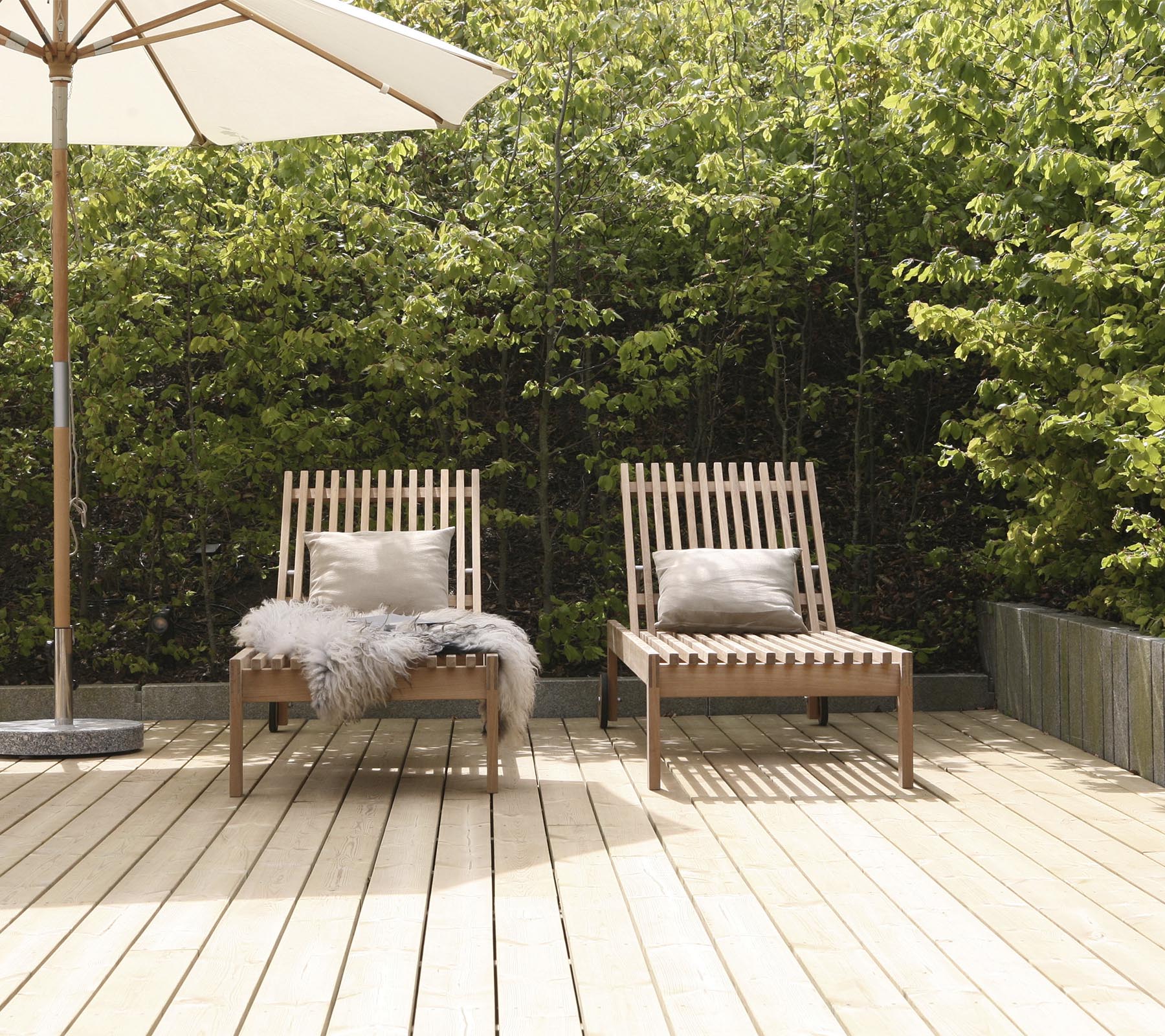 Dos sillas de estilo de madera con cojines, situadas en una terraza de madera, rodeadas de vegetación y con una gran sombrilla cerca.