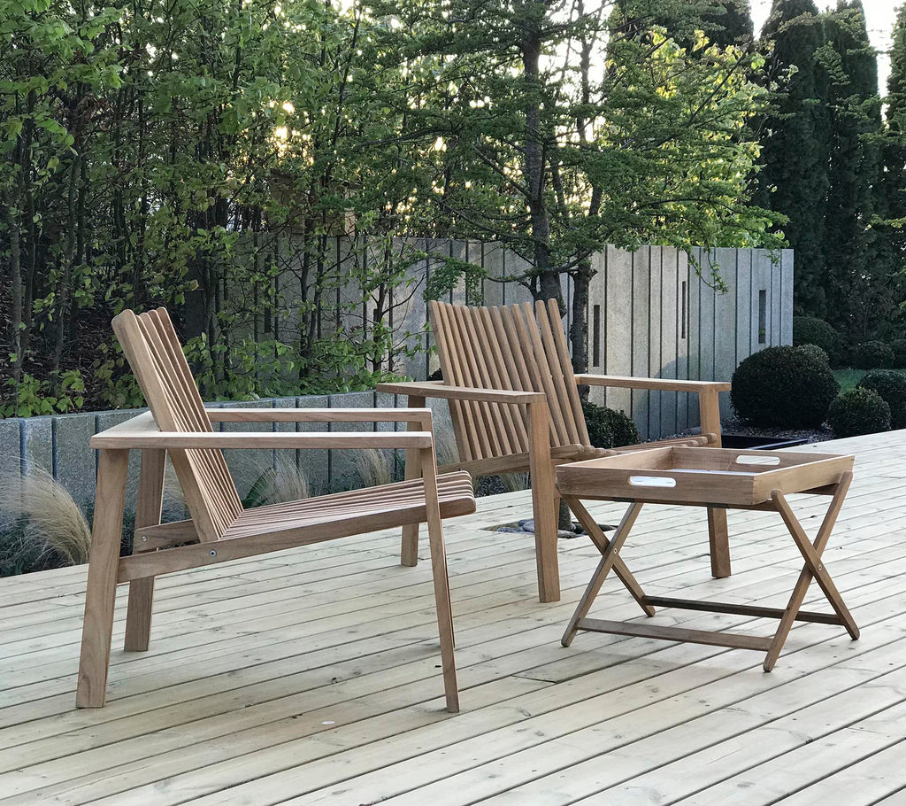 Dos sillas de madera y una pequeña mesa plegable, situadas frente a un fondo de vegetación y paneles de madera. Asientos al aire libre naturales y acogedores.