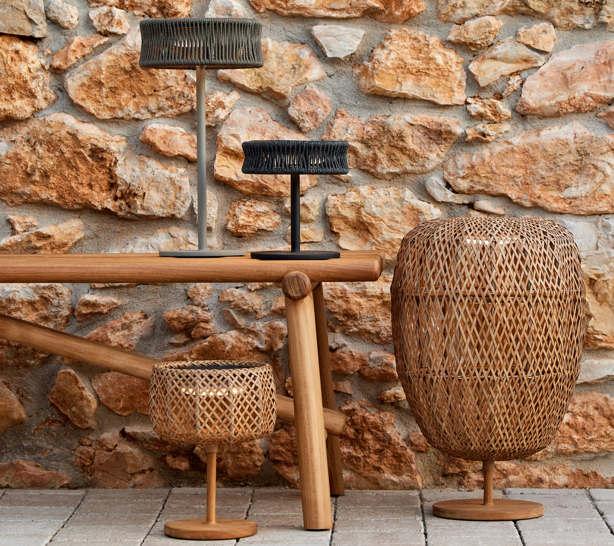 Tres lámparas decorativas de formas y tamaños variados sobre una mesa de madera, contra una pared de piedra texturizada.