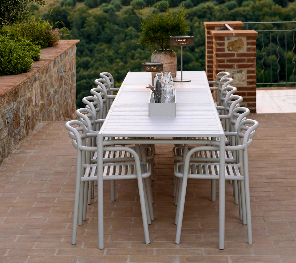 Una larga mesa blanca rodeada de varias sillas ligeras y elegantes, con elementos decorativos y vegetación que realzan el estilo del ambiente.