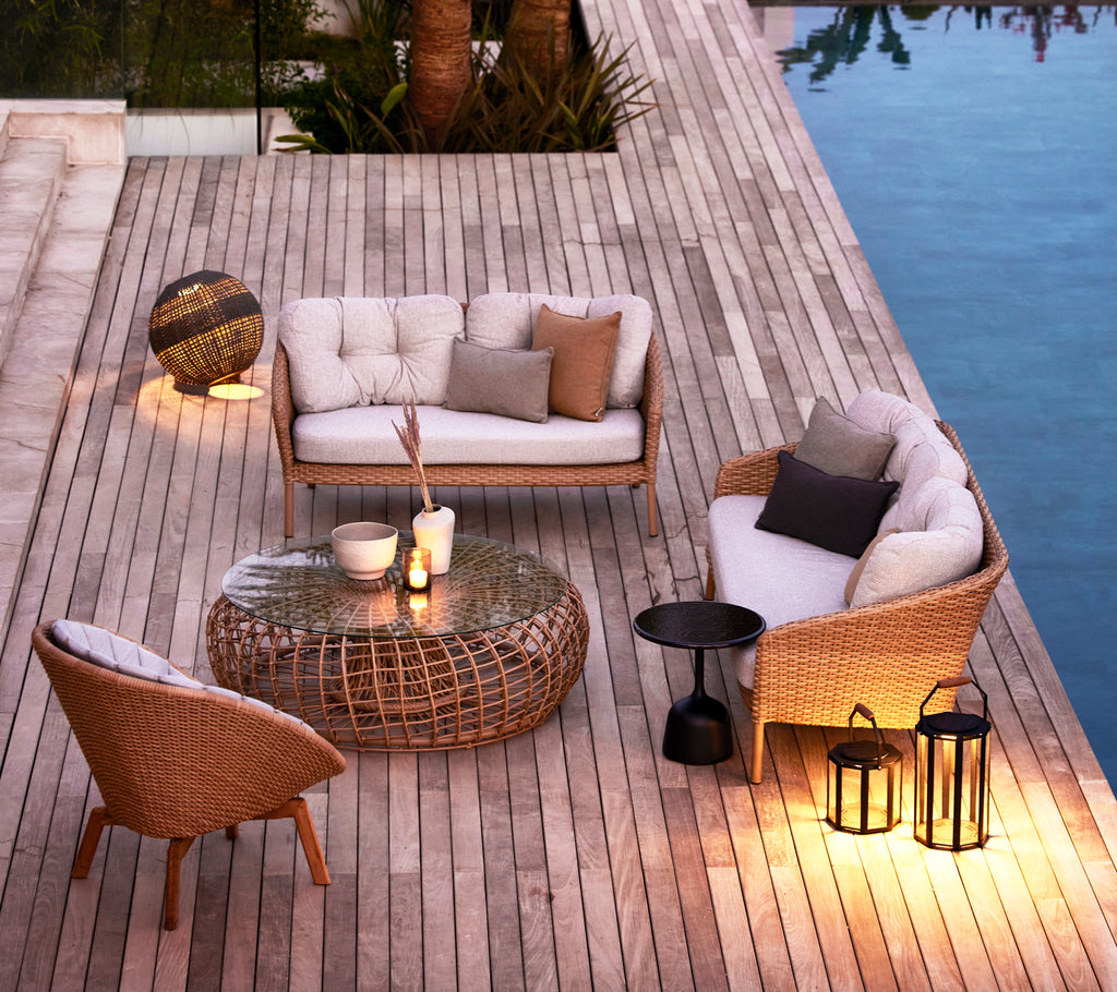 Área de estar elegante con muebles trenzados, una mesa de vidrio y lámparas decorativas, rodeada de madera y agua reflejante.