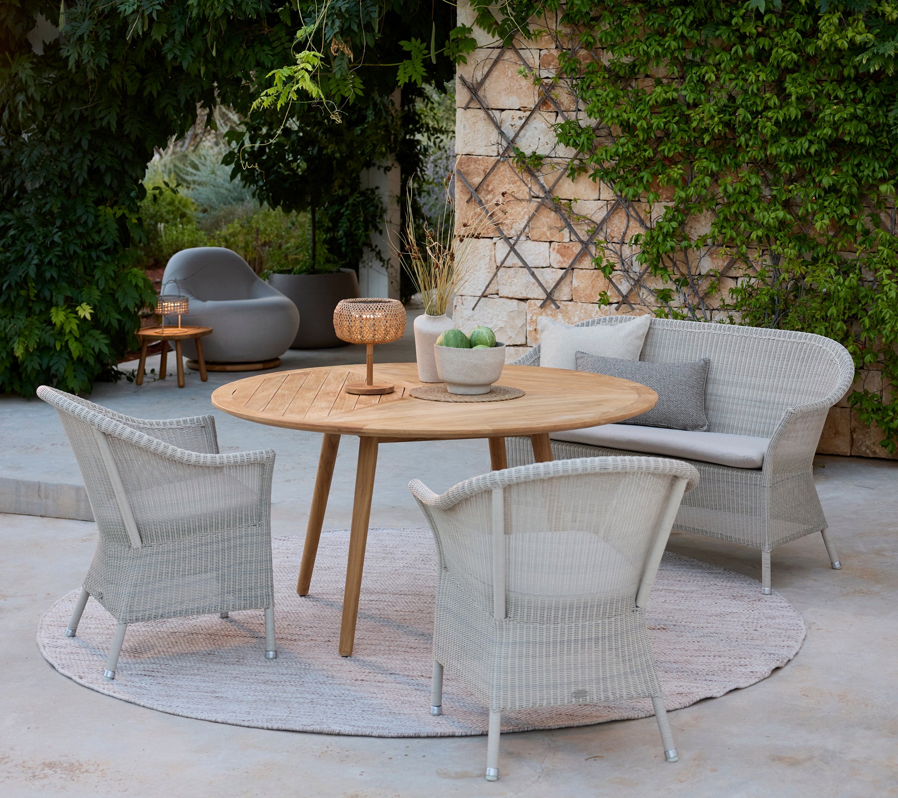 Zona de estar moderna con sillas grises, una mesa de madera redonda y un sofá gris rodeado de vegetación.
