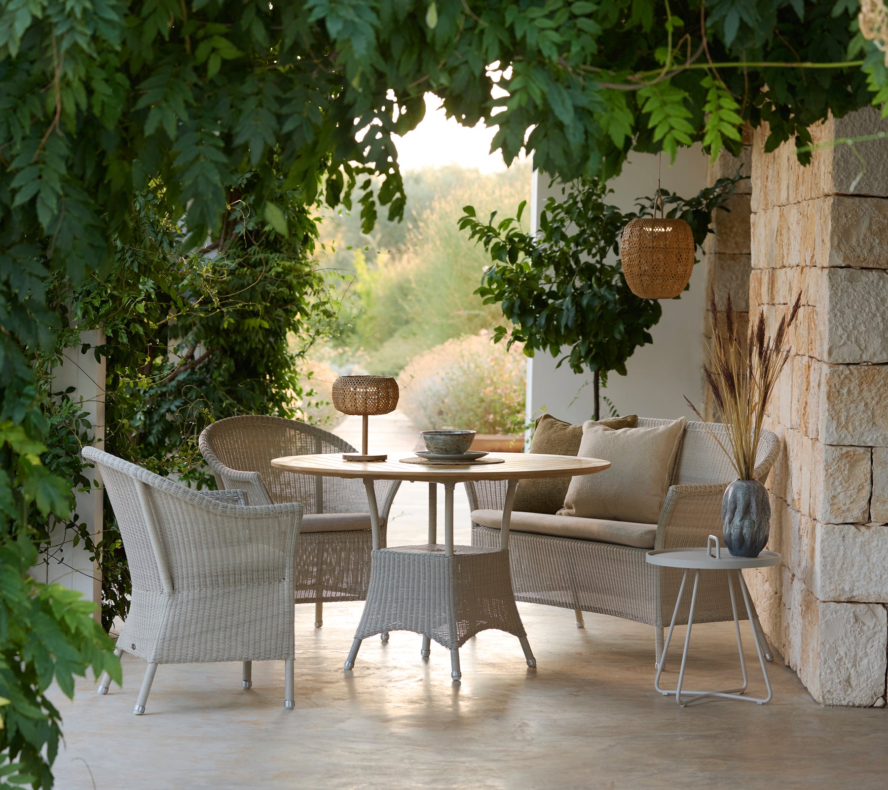 Conjunto de muebles de salón contemporáneos con sillas tejidas, mesa redonda y cojines en un entorno exterior sereno.
