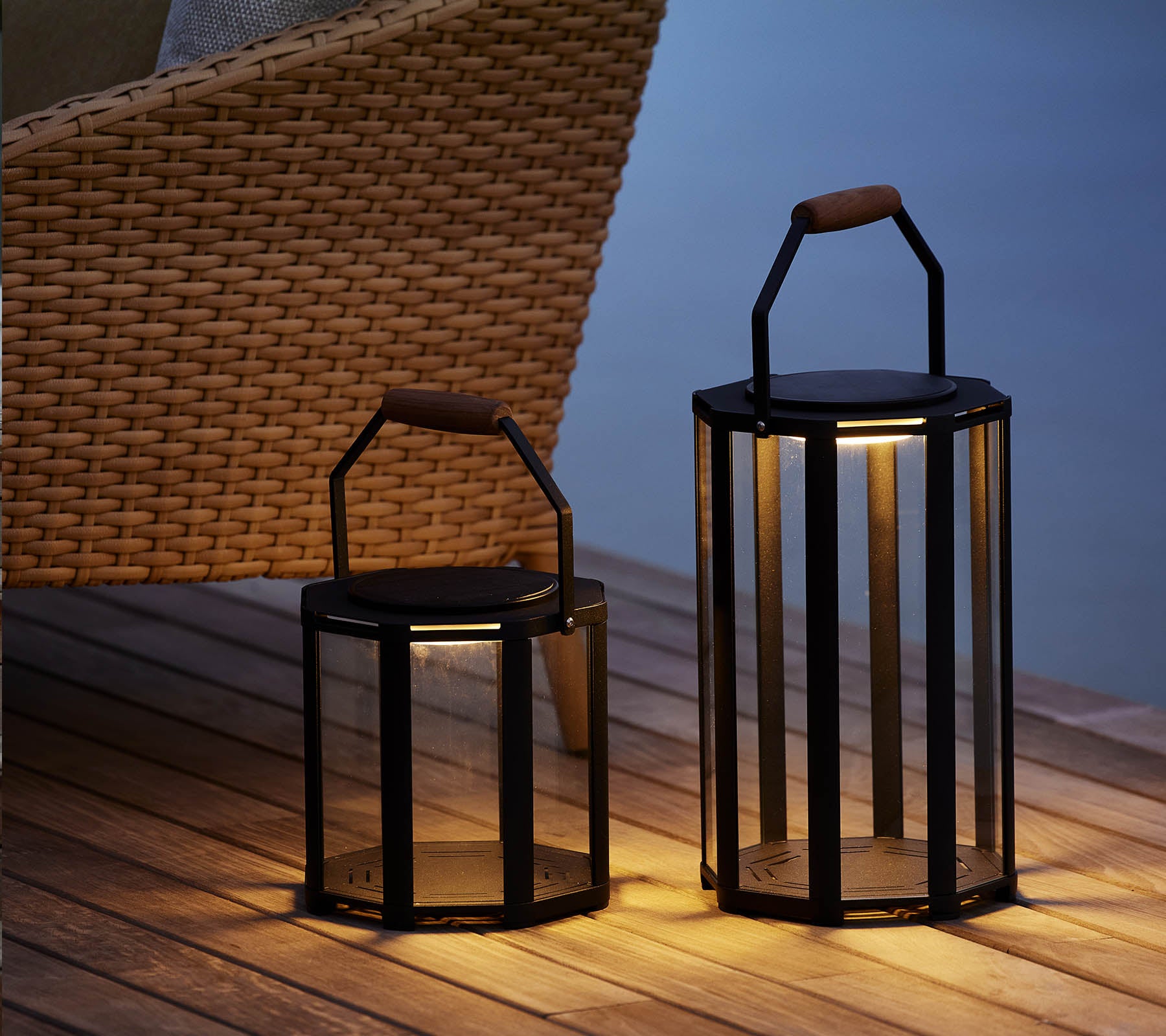 Dos elegantes faroles Lightluxe con paneles de vidrio y asas, que emiten un cálido resplandor, colocadas al lado de una silla trenzada.