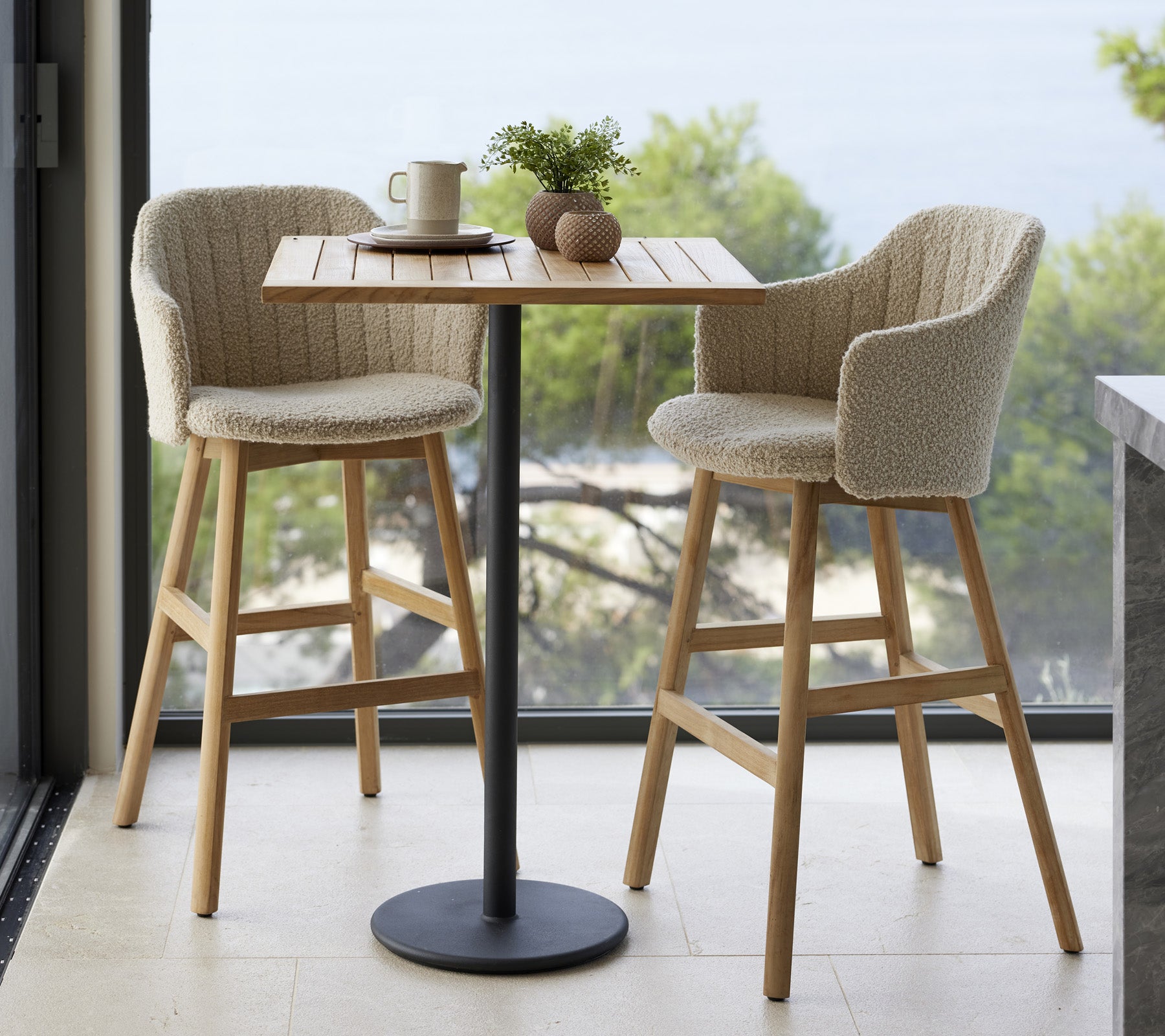 Una mesa pequeña con dos sillas tapizadas, una taza y elementos decorativos, colocada frente a un fondo natural.