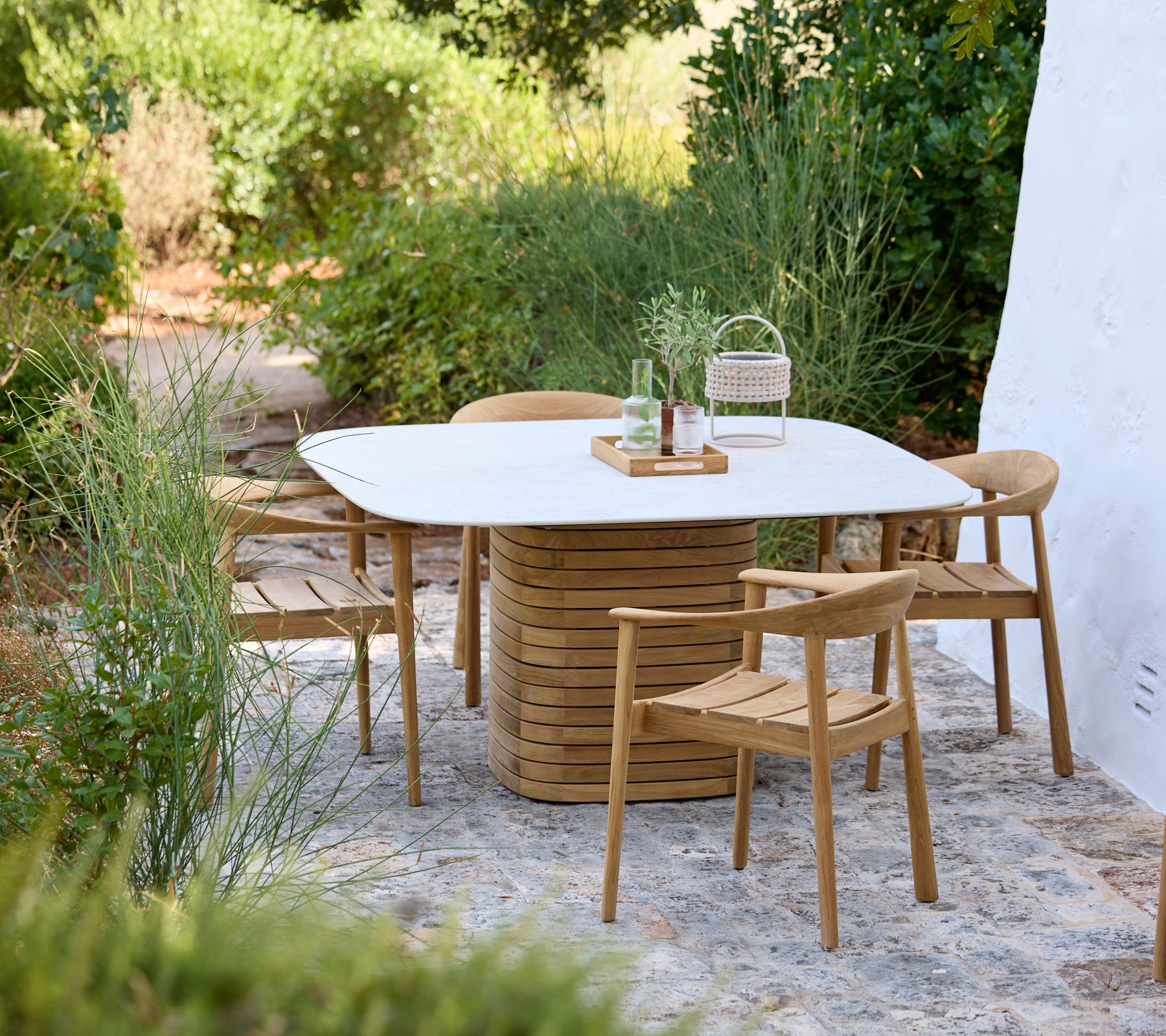 Configuración exterior moderna con una mesa blanca y sillas de madera rodeadas de vegetación.