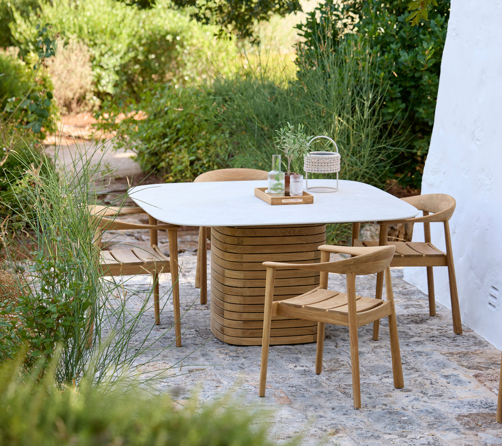 Configuración exterior moderna con una mesa blanca y sillas de madera rodeadas de vegetación.