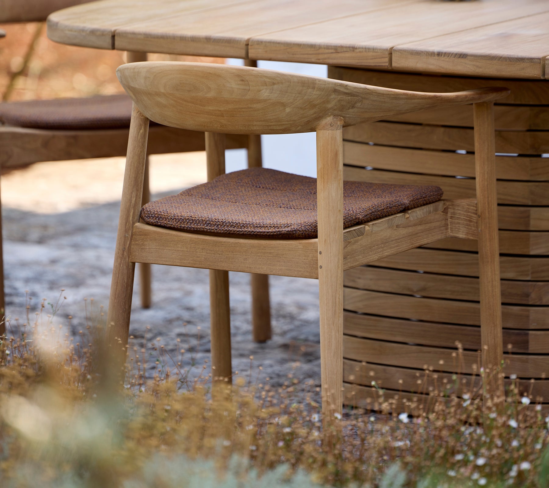 Silla de madera con cojín marrón al lado de una mesa redonda en un entorno al aire libre.