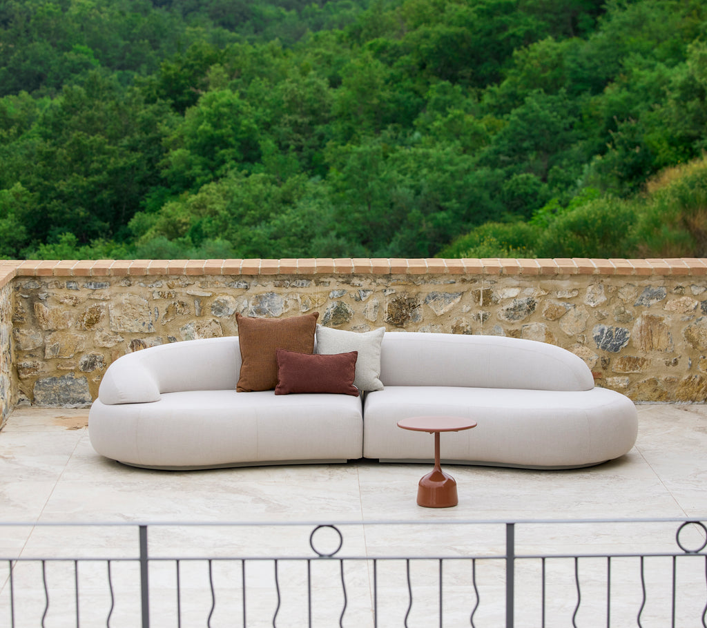 Un sofá moderno y curvado con tapicería neutra y almohadas decorativas, complementado por una mesa auxiliar elegante en un entorno natural.