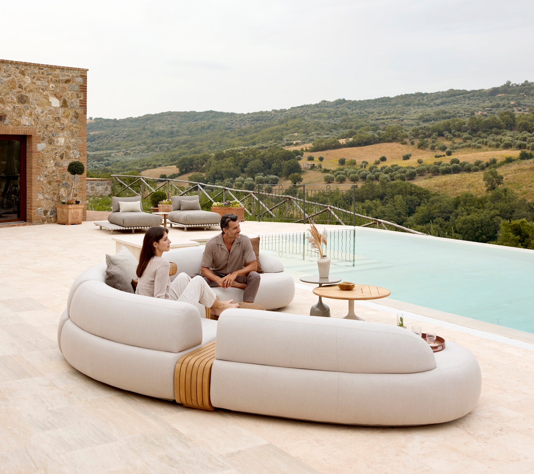 Una pareja se relaja en un sofá moderno y curvado junto a una mesa de café elegante, con una hermosa vista de colinas ondulantes en el fondo.