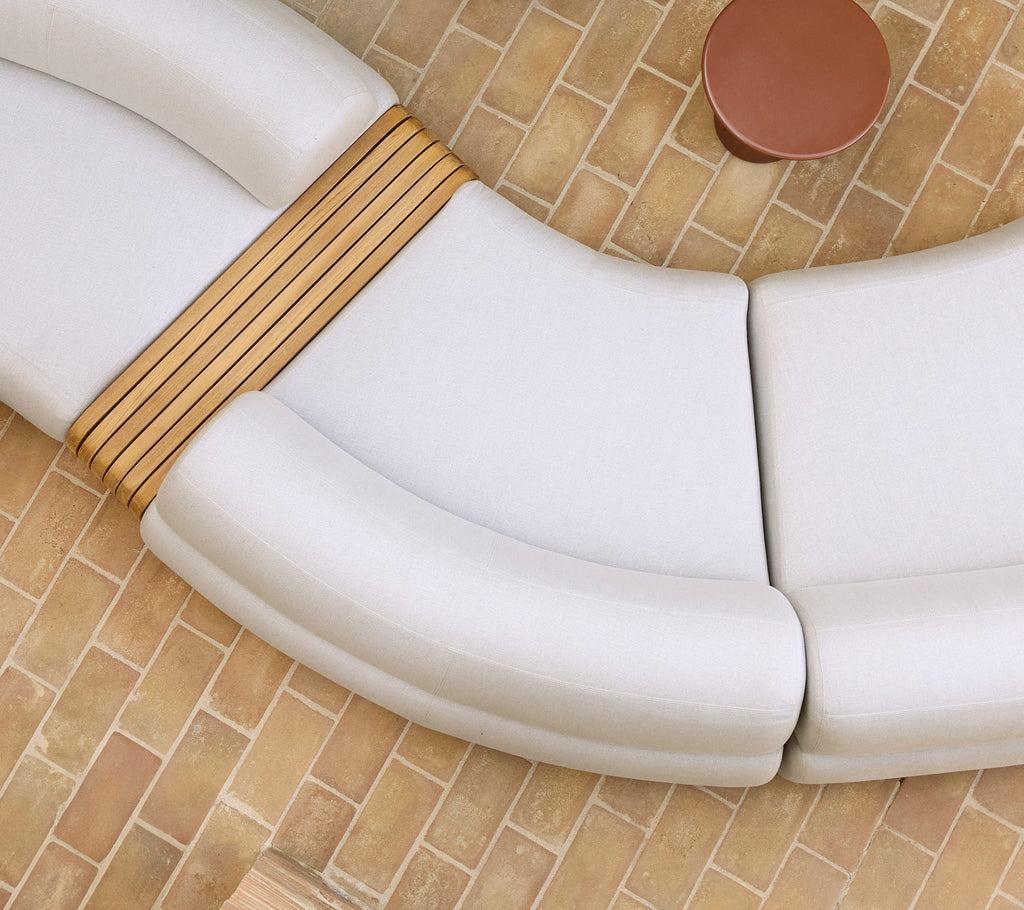 Módulo central de sofá suave con curvas suaves y acentos de madera al lado de una mesa de café redonda sobre un suelo texturizado.