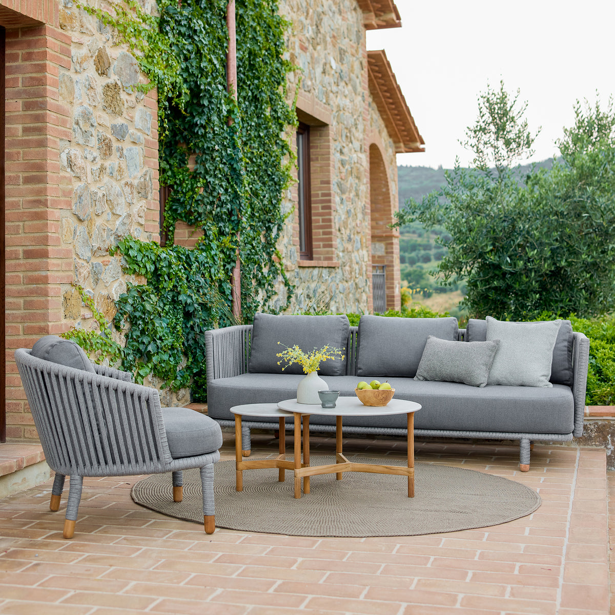 Juego de sofá gris moderno con sillón a juego y una mesa de café redonda, rodeado de vegetación y arquitectura de piedra rústica.