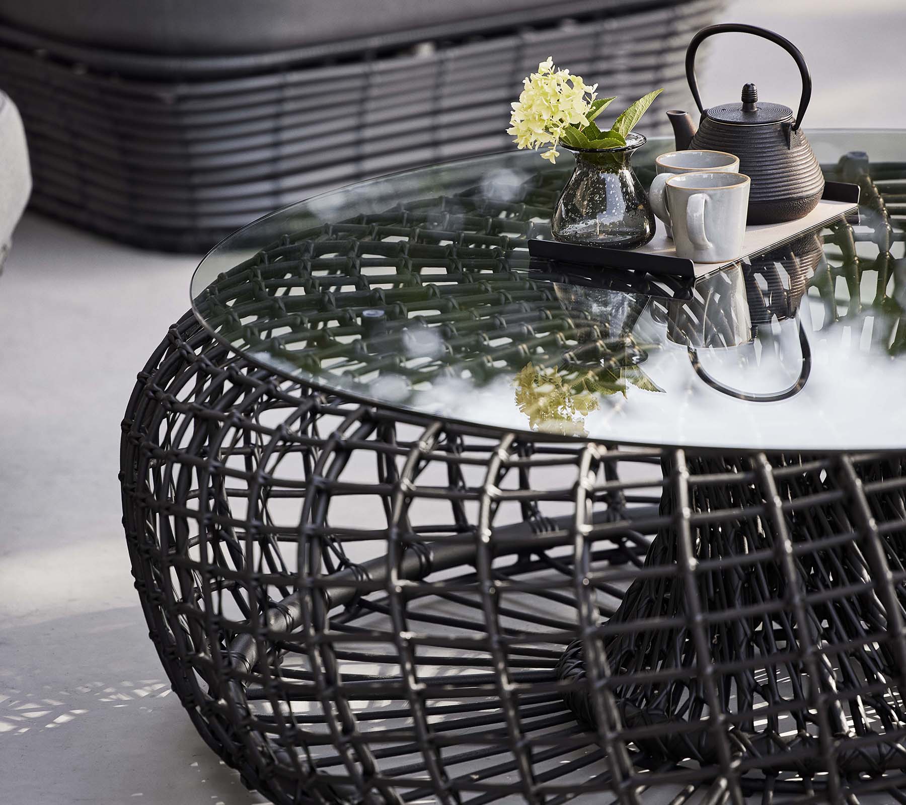 Mesa de café Nest con una base trenzada, una superficie de vidrio y un set de té con flores para un aspecto moderno y elegante.