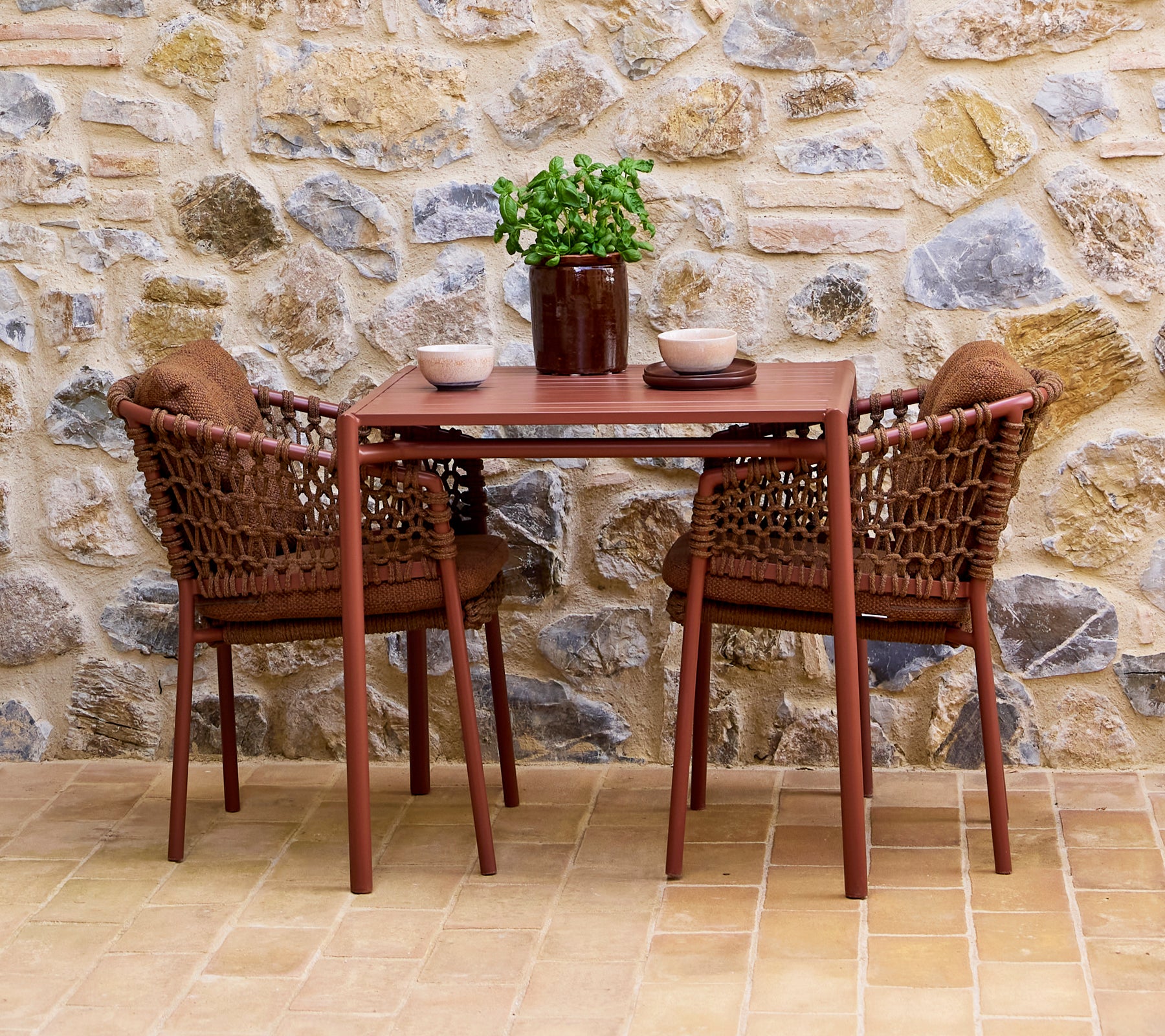 Sillas de umbra marrón combinadas con una mesa a juego, acentuadas por una planta en maceta y cuencos decorativos, contra un fondo de pared de piedra.