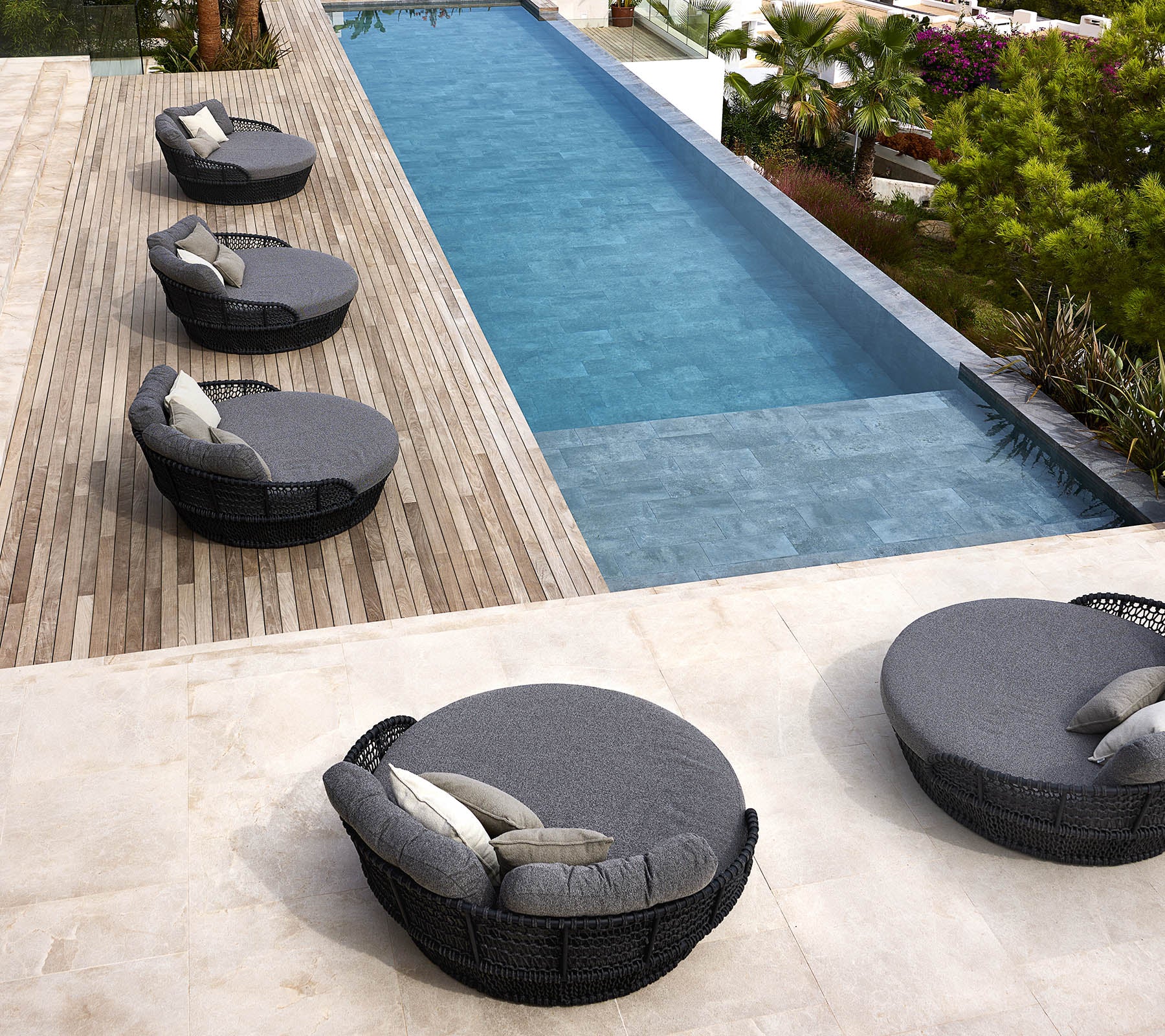 Cuatro camas redondas de día junto al océano con cojines están dispuestas junto a una elegante piscina rodeada de vegetación y terraza de madera.