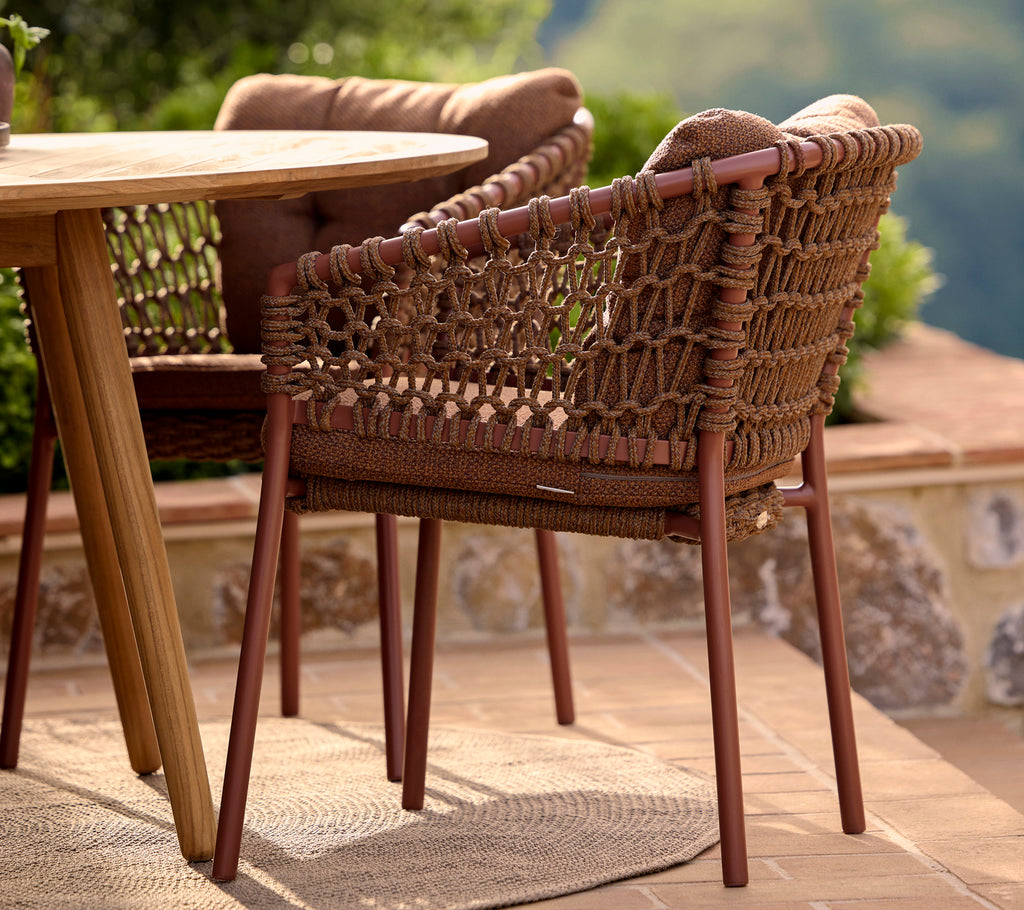Silla de comedor marrón ámbar con un diseño tejido y patas robustas, situada junto a una mesa redonda y rodeada de vegetación.