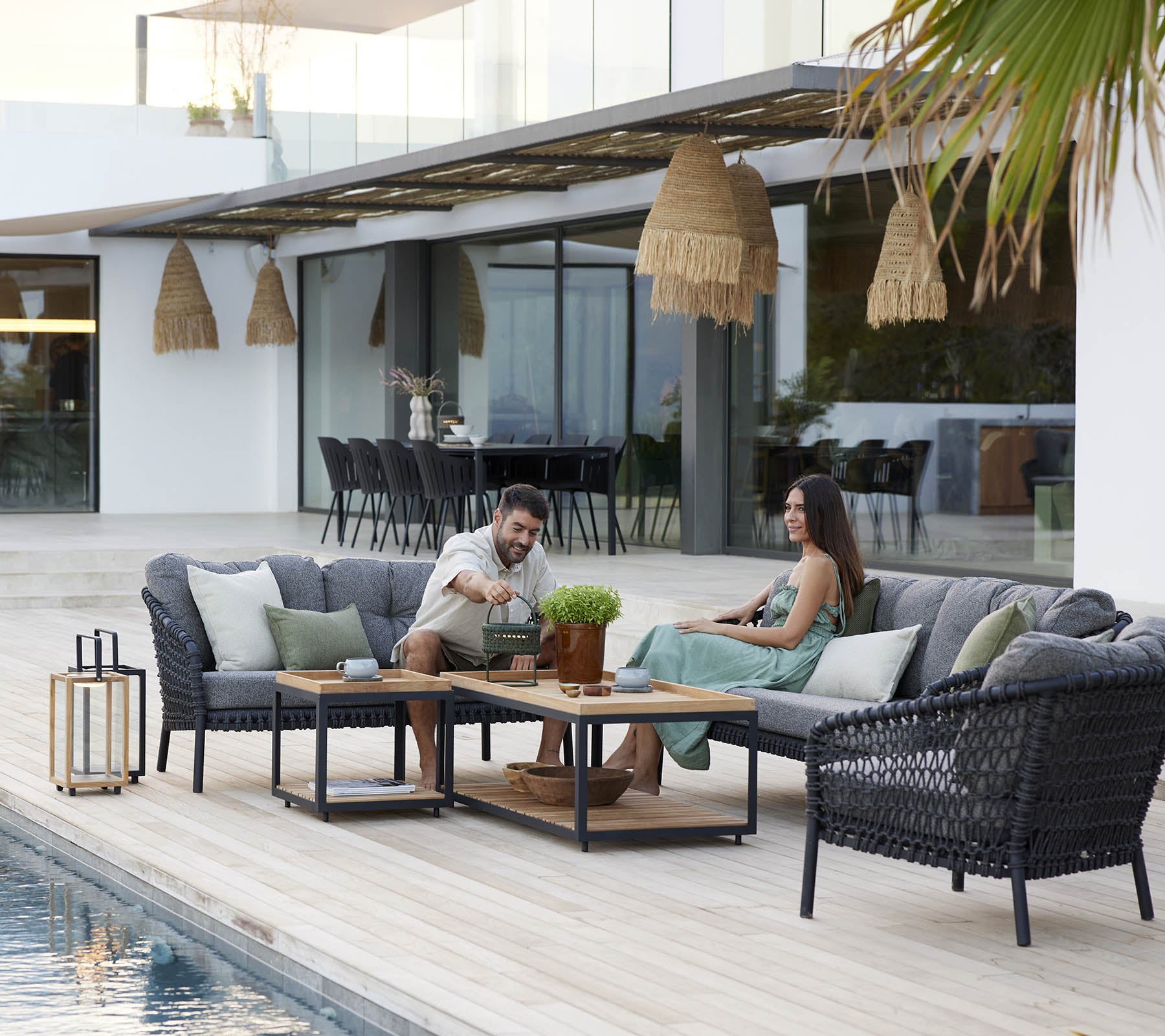 Una pareja disfrutando de su tiempo de ocio en un acogedor conjunto de salón, rodeado de plantas verdes y una decoración elegante, con una piscina serena cerca.