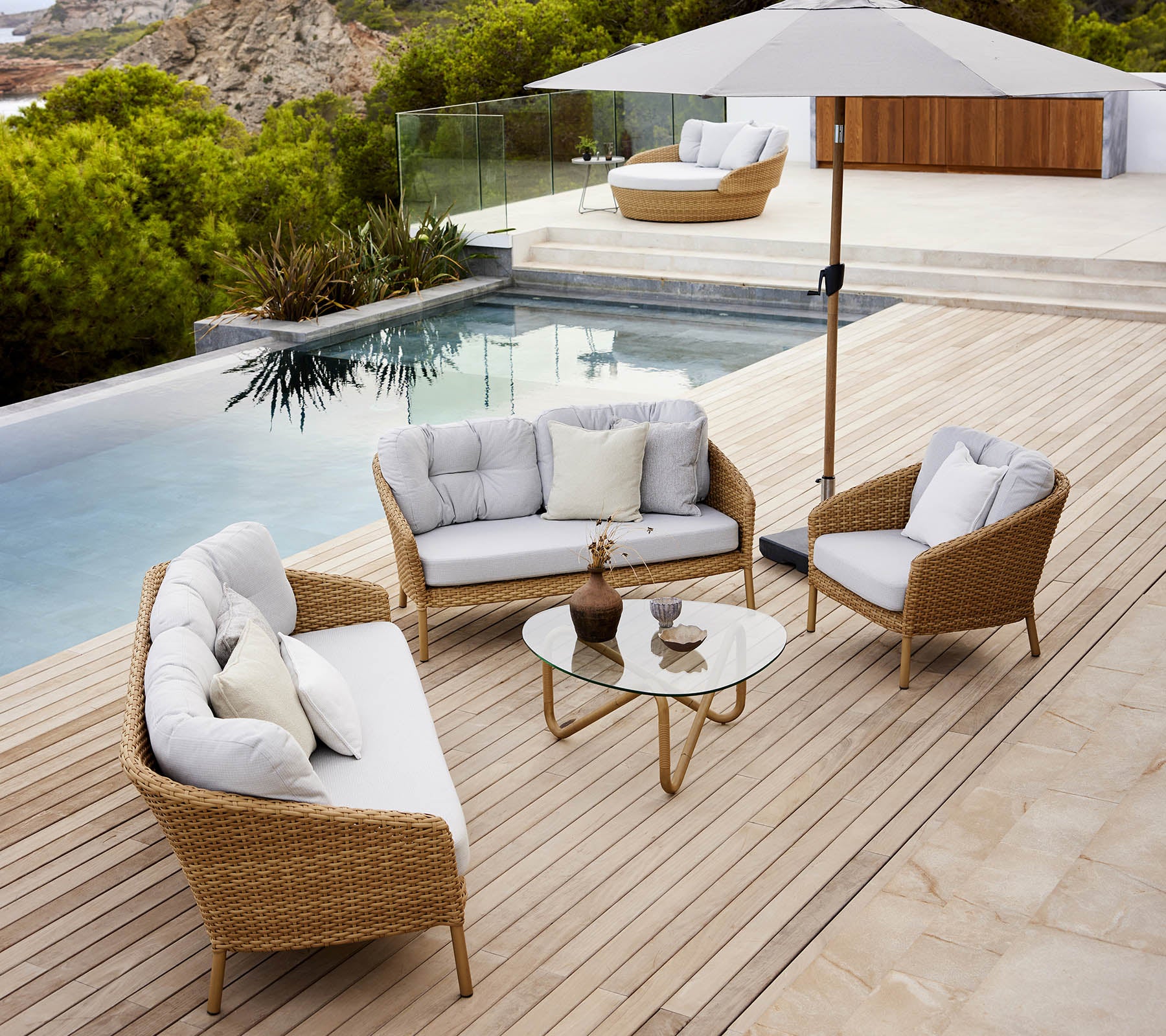 Moderna zona de lounge al aire libre con una mesa de vidrio, asientos cómodos y una sombrilla junto a la piscina con exuberante vegetación.