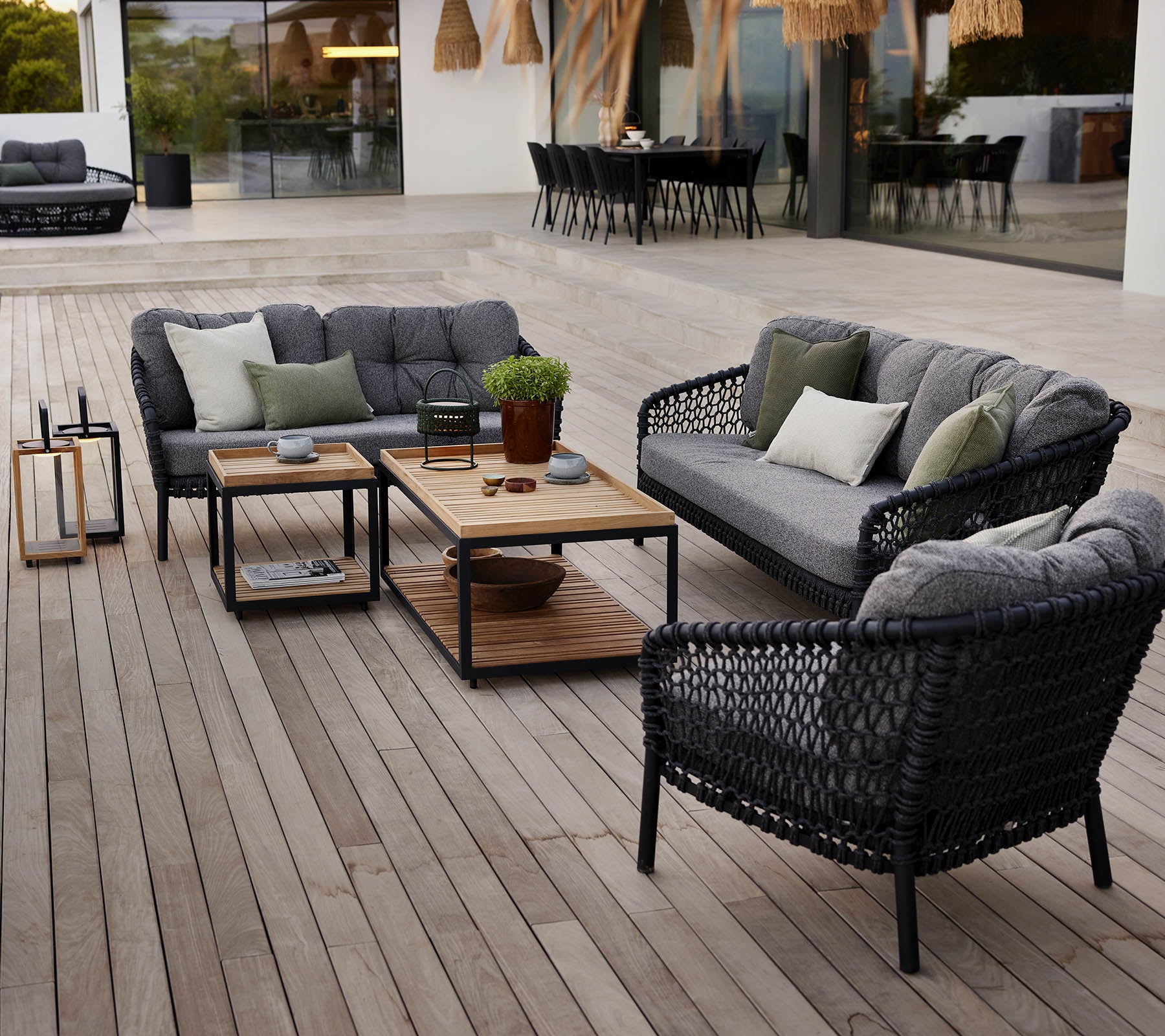 Área de lounge exterior elegante con muebles de ratán negros, cojines grises, una mesa de café de madera y plantas en macetas en una terraza de madera.