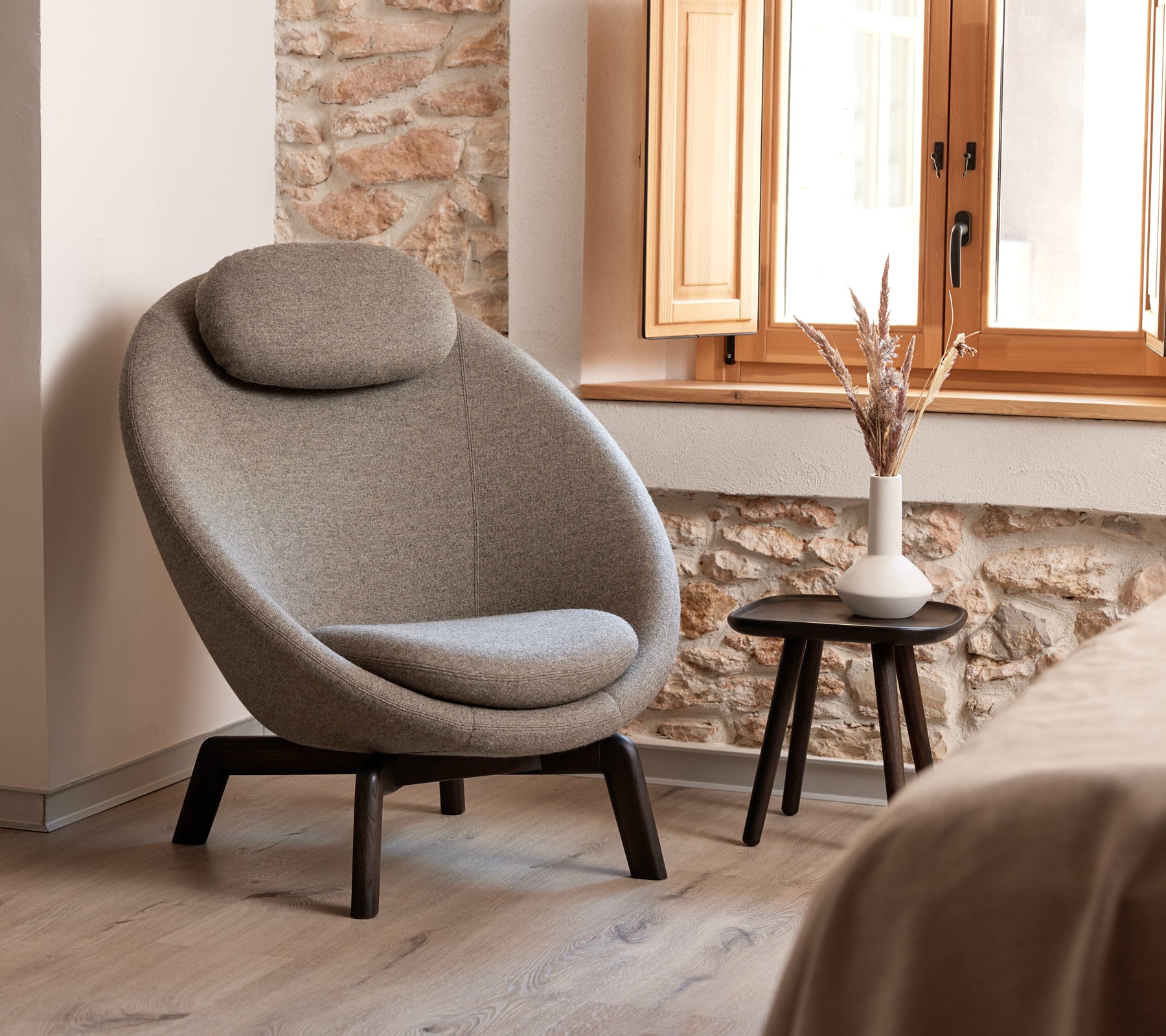 Silla tapizada elegante y curvada con un reposacabezas cómodo, acompañada de una pequeña mesa auxiliar con un jarrón y elementos decorativos.