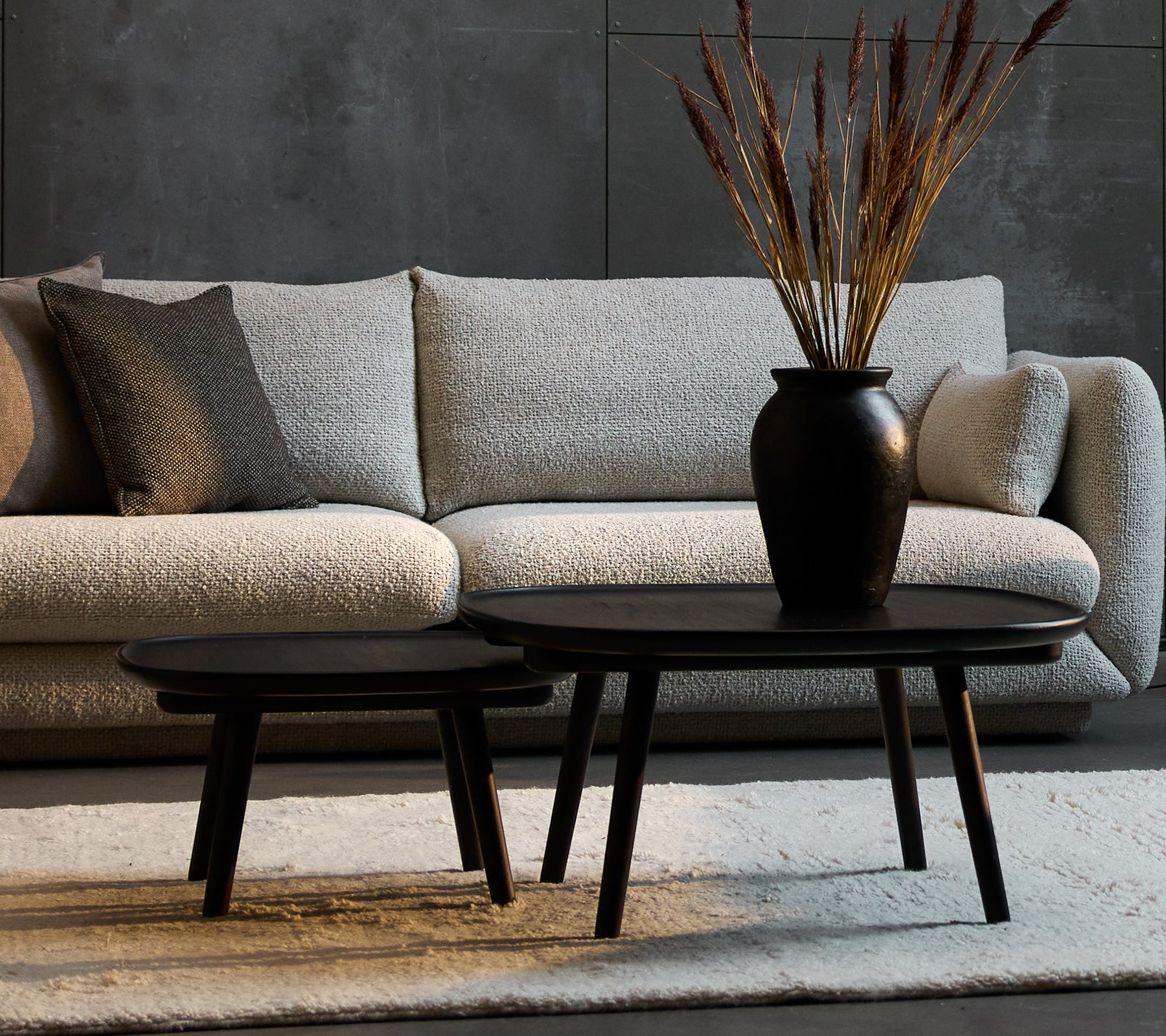 Dos elegantes mesas de café negras de diferentes tamaños, complementadas con un jarrón decorativo con plantas secas, colocadas frente a un sofá acogedor.