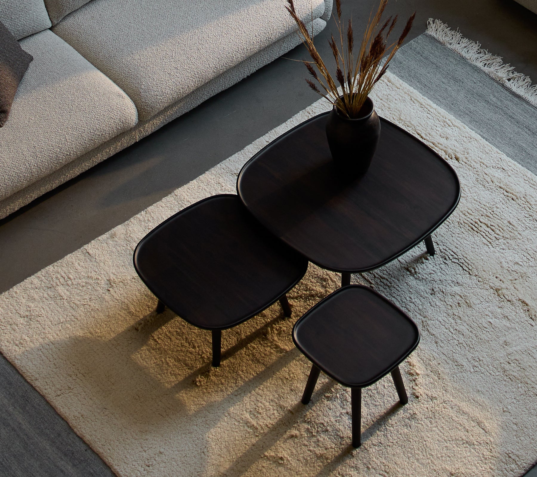 Tres mesas de café Pace elegantes de diferentes tamaños, con un diseño elegante y un acabado oscuro, acompañadas de un jarrón decorativo y plantas secas.