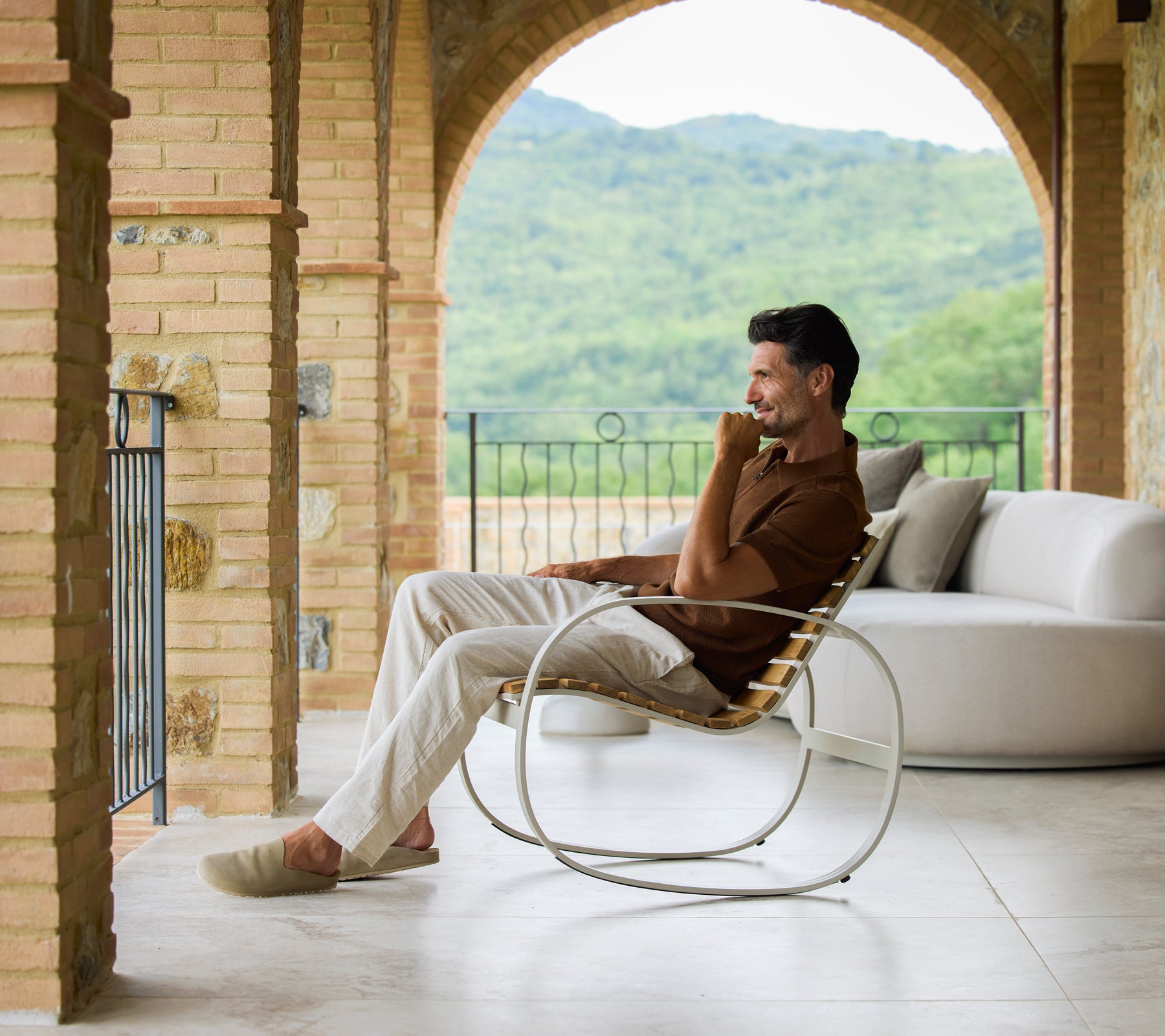 Hombre relajándose en una silla mecedora de color arena, disfrutando de vistas escénicas y de una atmósfera serena. Tonos suaves y neutros destacan la comodidad.
