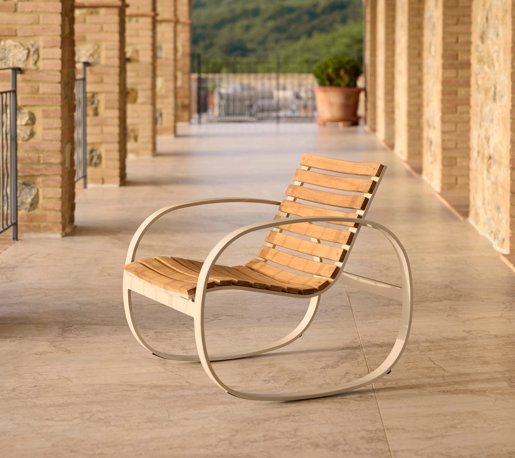 Silla mecedora Parc con estructura de aluminio y listones de madera, situada en un patio con suelo de piedra y un fondo escénico.