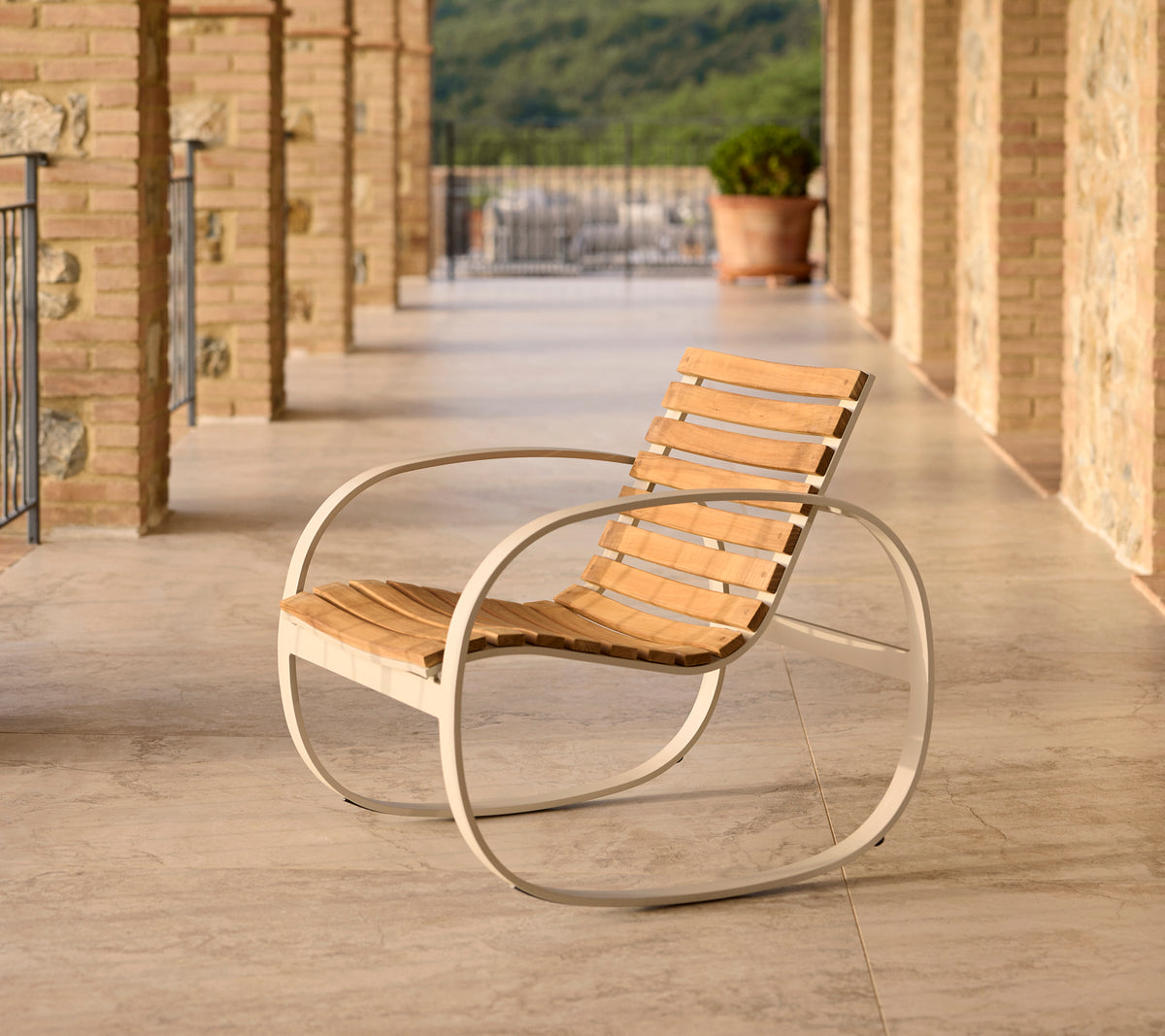 Silla mecedora Parc con estructura de aluminio y listones de madera, situada en un patio con suelo de piedra y un fondo escénico.