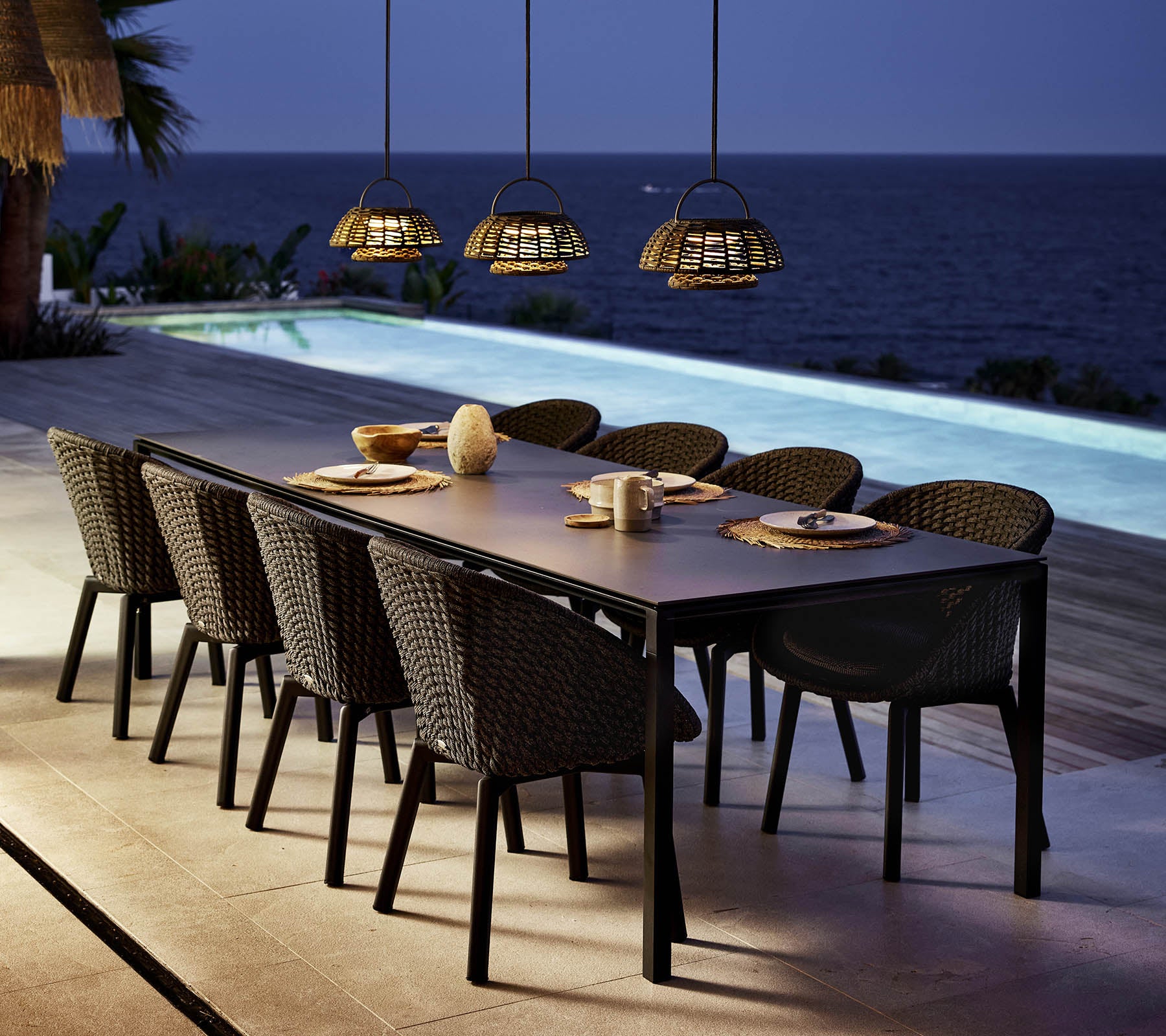 Una elegante disposición de comedor presenta una larga mesa rodeada de sillas de pavo real, iluminada por luces colgantes sobre una serena vista del agua.