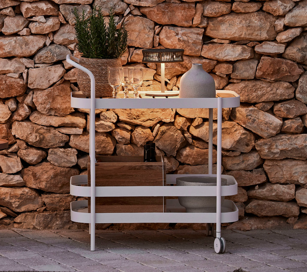 Un carrito elegante con una parte superior de madera, estructura blanca, maceta decorativa y una iluminación elegante contra un fondo de pared de piedra.