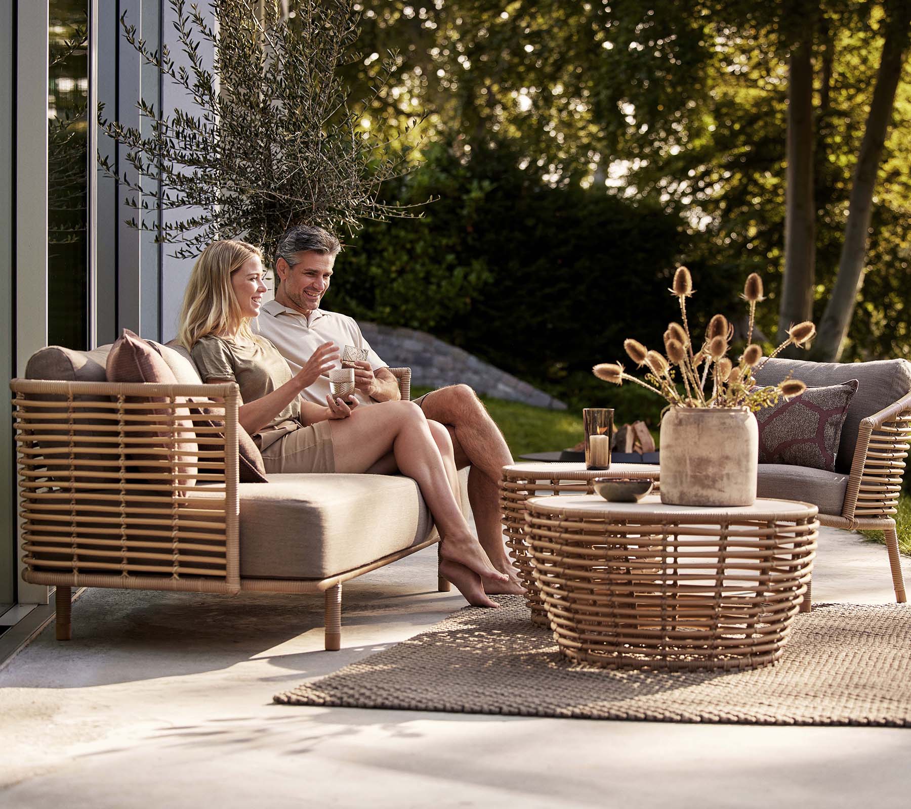 Una pareja disfruta de bebidas en un sofá Sense y una silla de lounge, acompañados de una elegante mesa de café en forma de cesta y un jarrón decorativo.