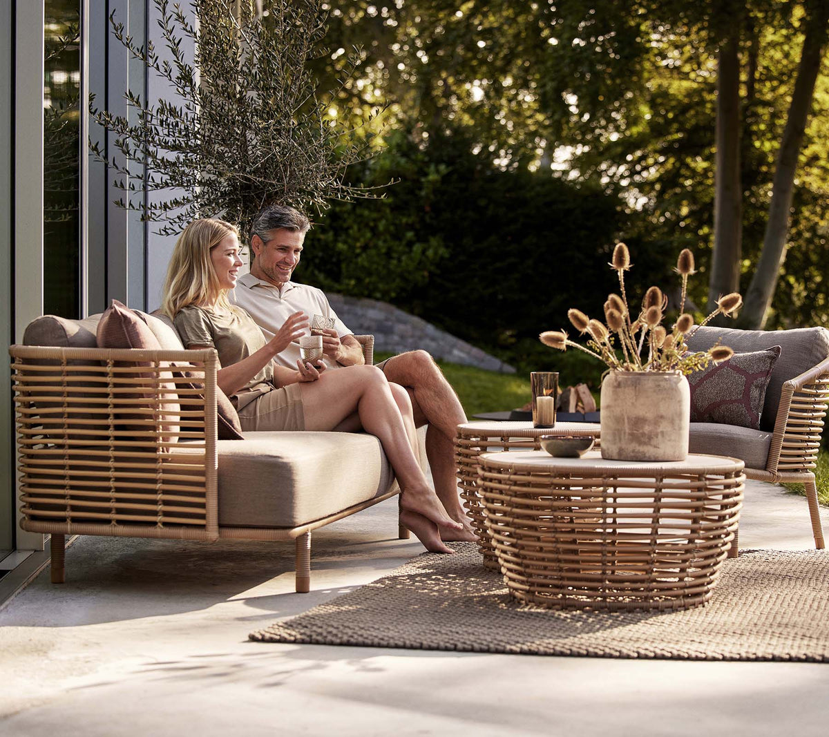 Una pareja disfruta de bebidas en un sofá Sense y una silla de lounge, acompañados de una elegante mesa de café en forma de cesta y un jarrón decorativo.