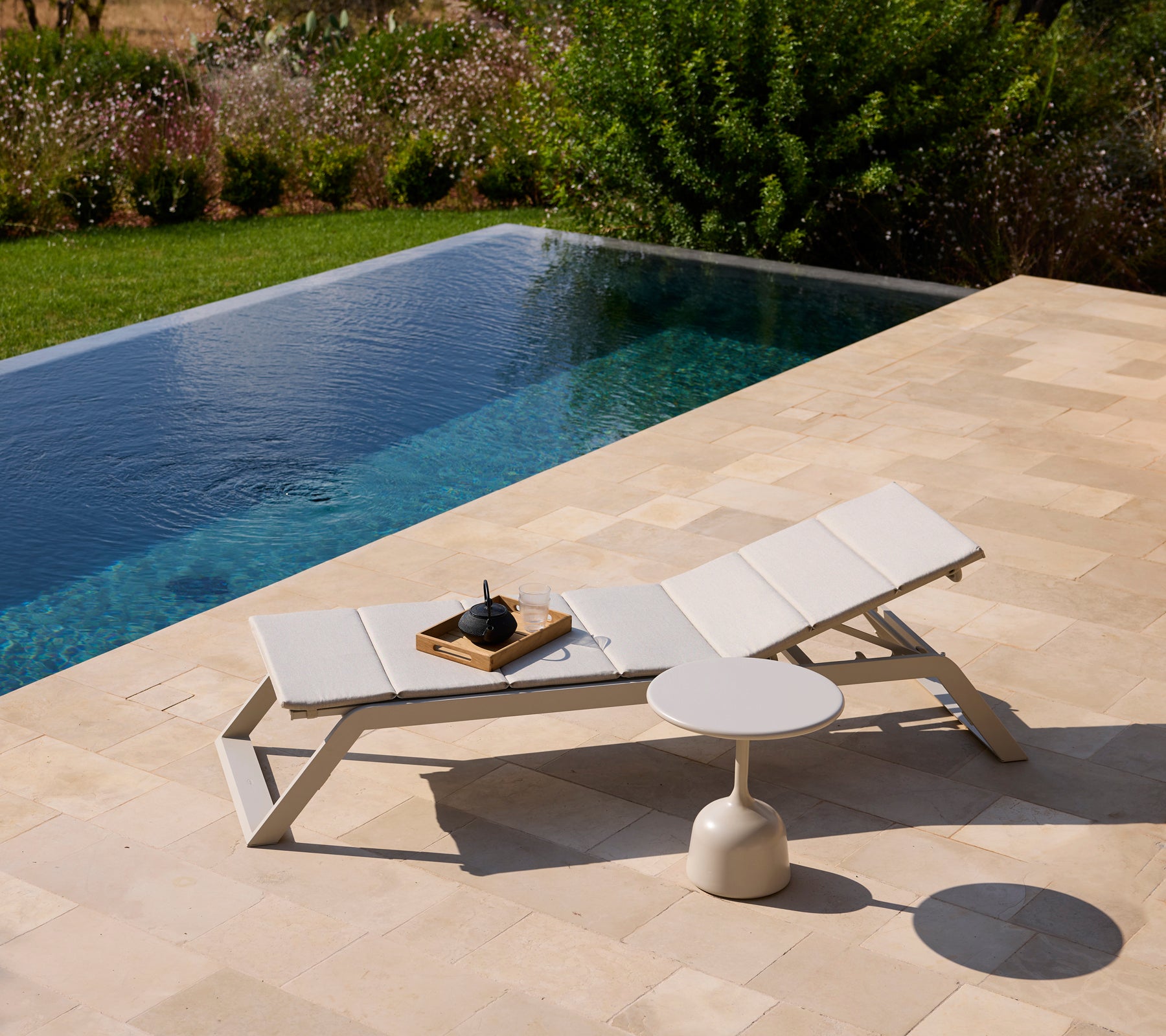 Silla lounge blanca moderna y mesa auxiliar junto a una piscina rodeada de vegetación.