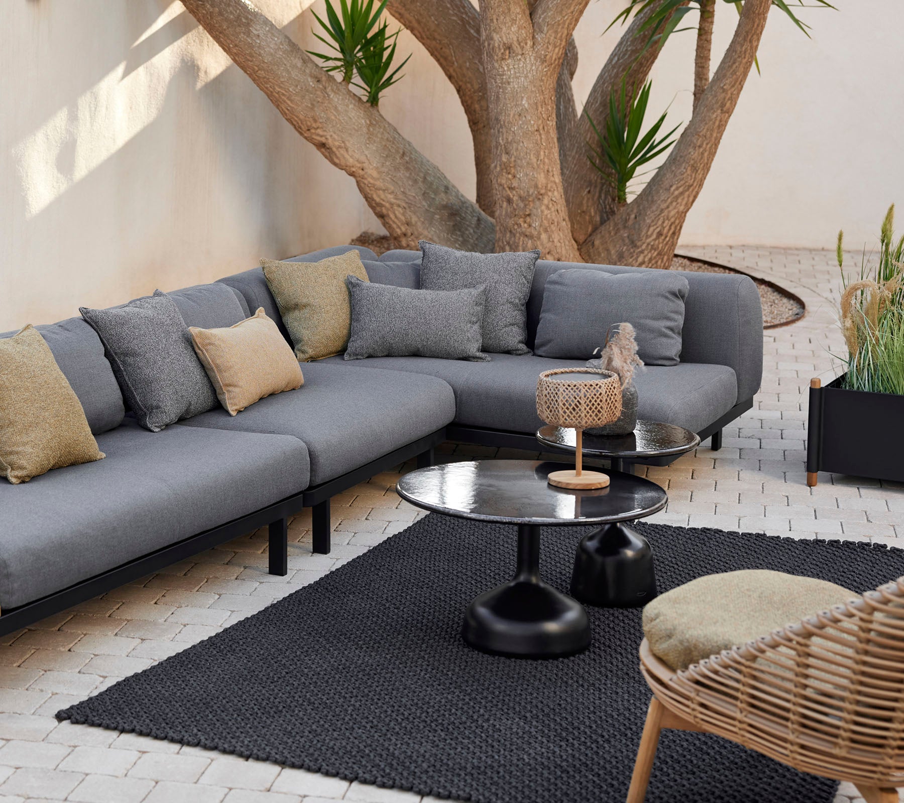 Zona de estar con un sofá modular gris con almohadas decorativas, una mesa de café elegante y una alfombra texturizada rodeada de vegetación.
