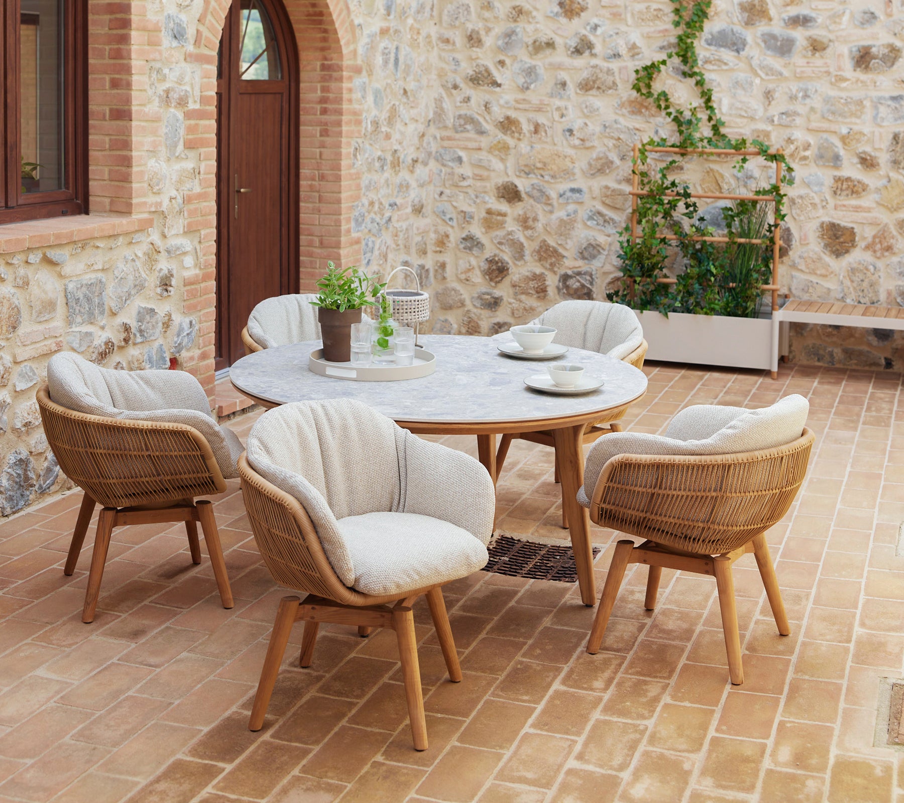 Una elegante disposición de comedor con una mesa redonda rodeada de sillas giratorias con patas de teca y cómodos cojines, complementada con vegetación.