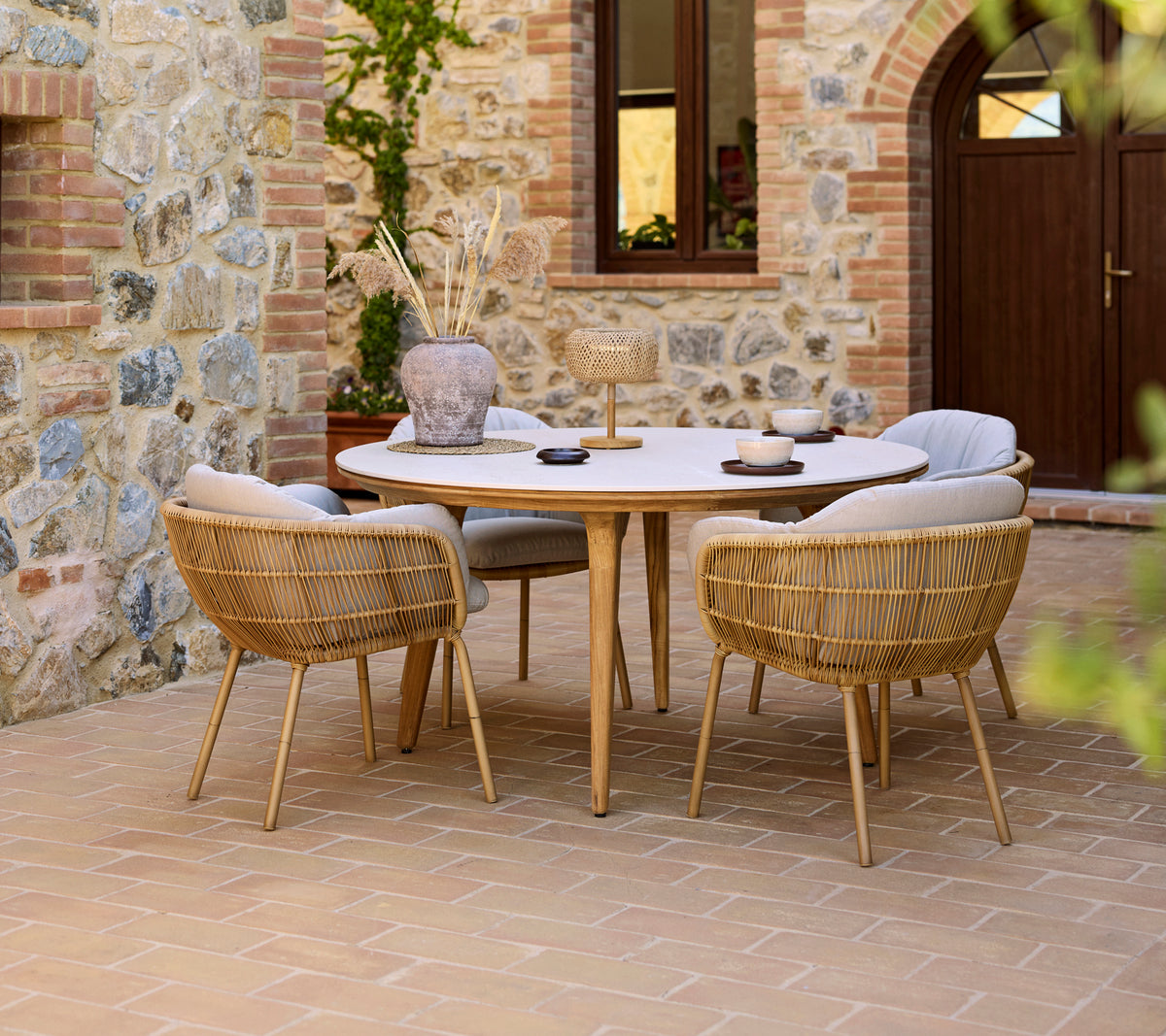 Montaje de comedor al aire libre con una mesa redonda Aspect y sillas Stay, con un fondo de pared de piedra.