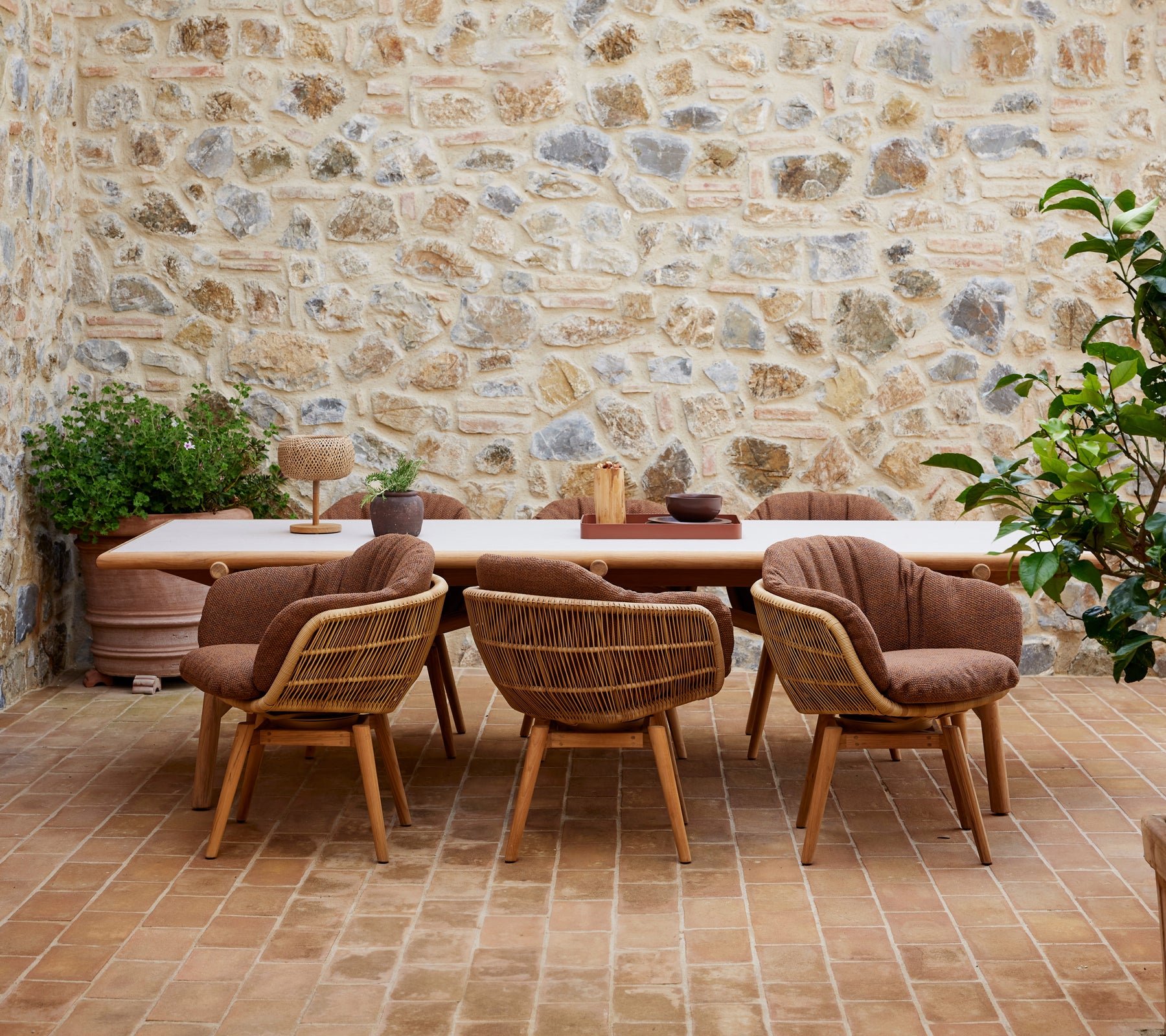 Una mesa de comedor de madera rodeada de sillas elegantes con patas de teca, contra una pared de piedra texturizada y adornada con plantas verdes.