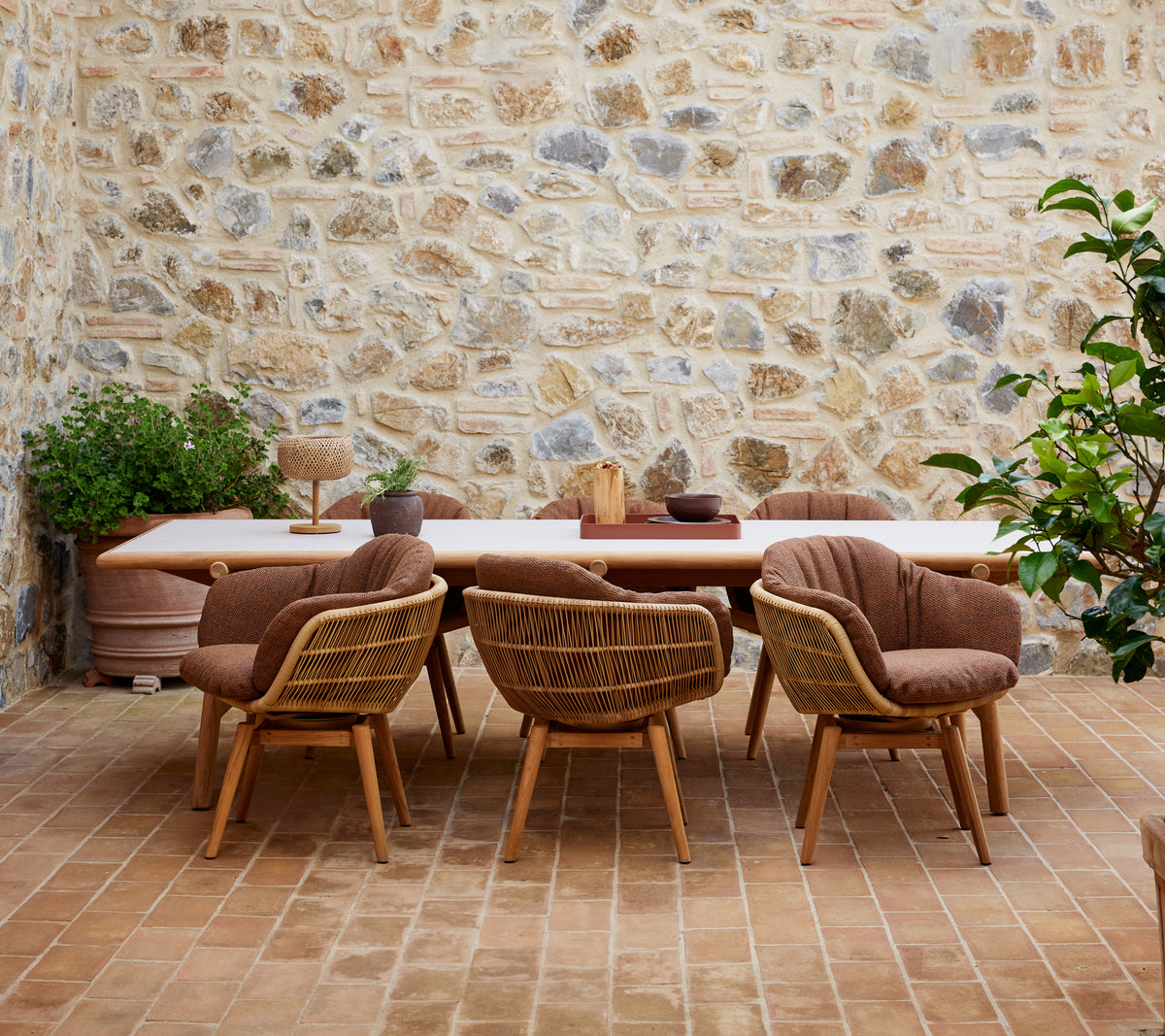 Una mesa de comedor de madera rodeada de sillas elegantes con patas de teca, contra una pared de piedra texturizada y adornada con plantas verdes.